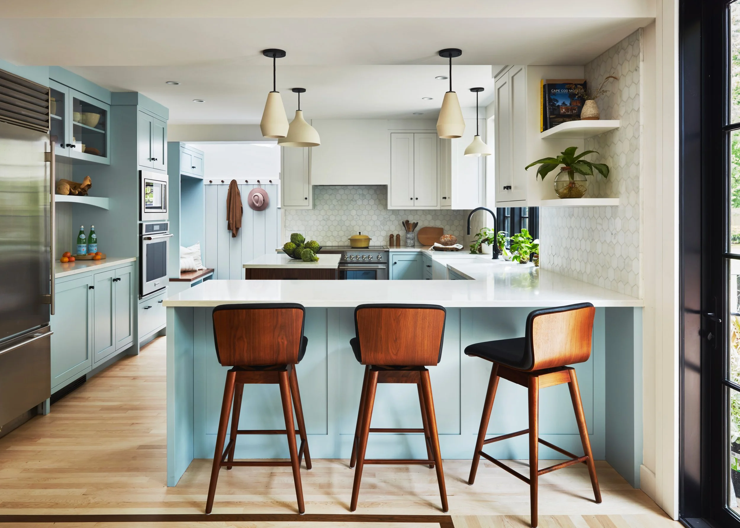 Bright kitchen with light blue cabinets, white countertops, and wooden stools at a breakfast bar. Pendant lights hang from the ceiling, and there are shelves with plants and books on the wall.