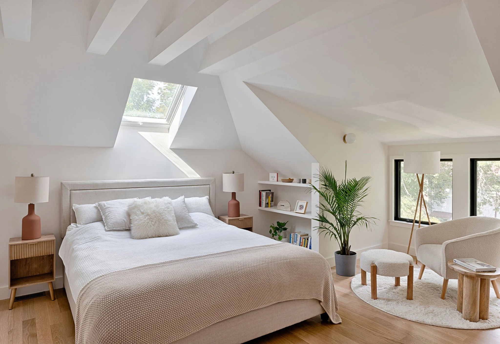 A cozy bedroom with a sloped ceiling, skylight window, and minimalist decor. The bed has a white headboard, white bedding, and fluffy pillows. Two bedside tables with lamps, an open bookshelf, a tall floor lamp, a potted plant, a white armchair, and a small round rug complete the room.