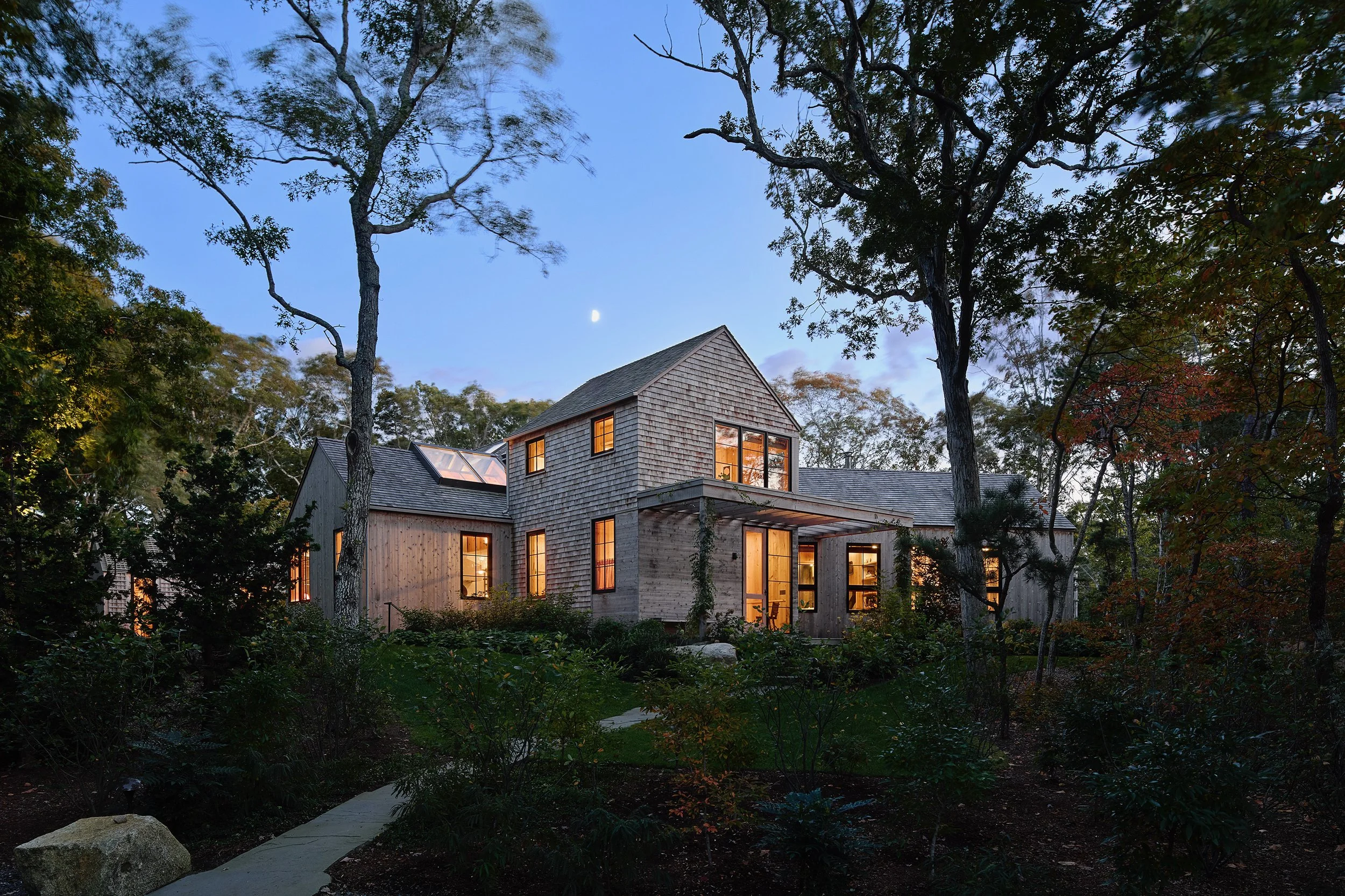 A modern house surrounded by trees at dusk, with lights on inside and a clear sky showing the moon.