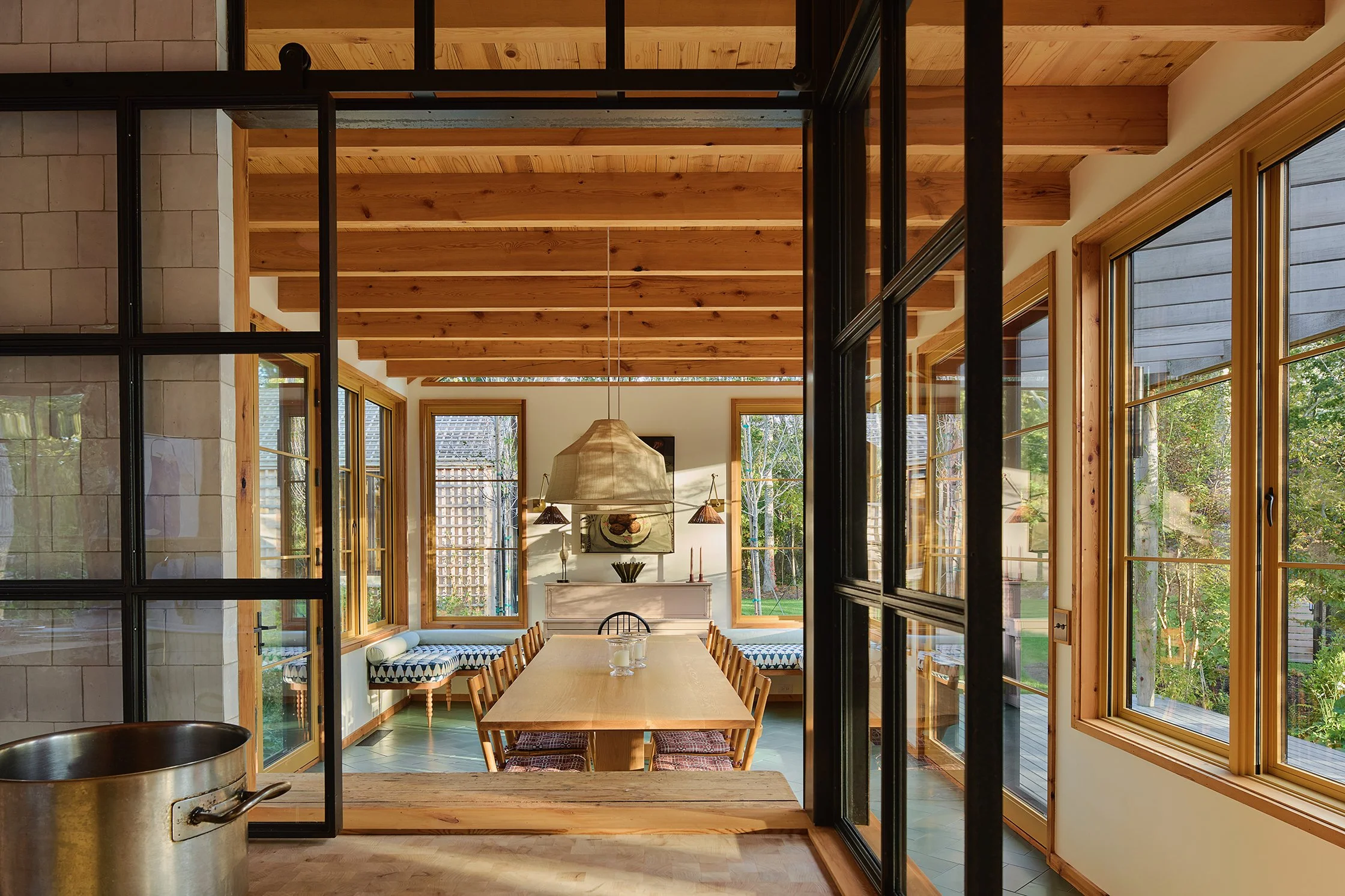 View of a cozy dining area with a wooden table and chairs, large windows with a balcony and greenery outside, in a warm, modern home.