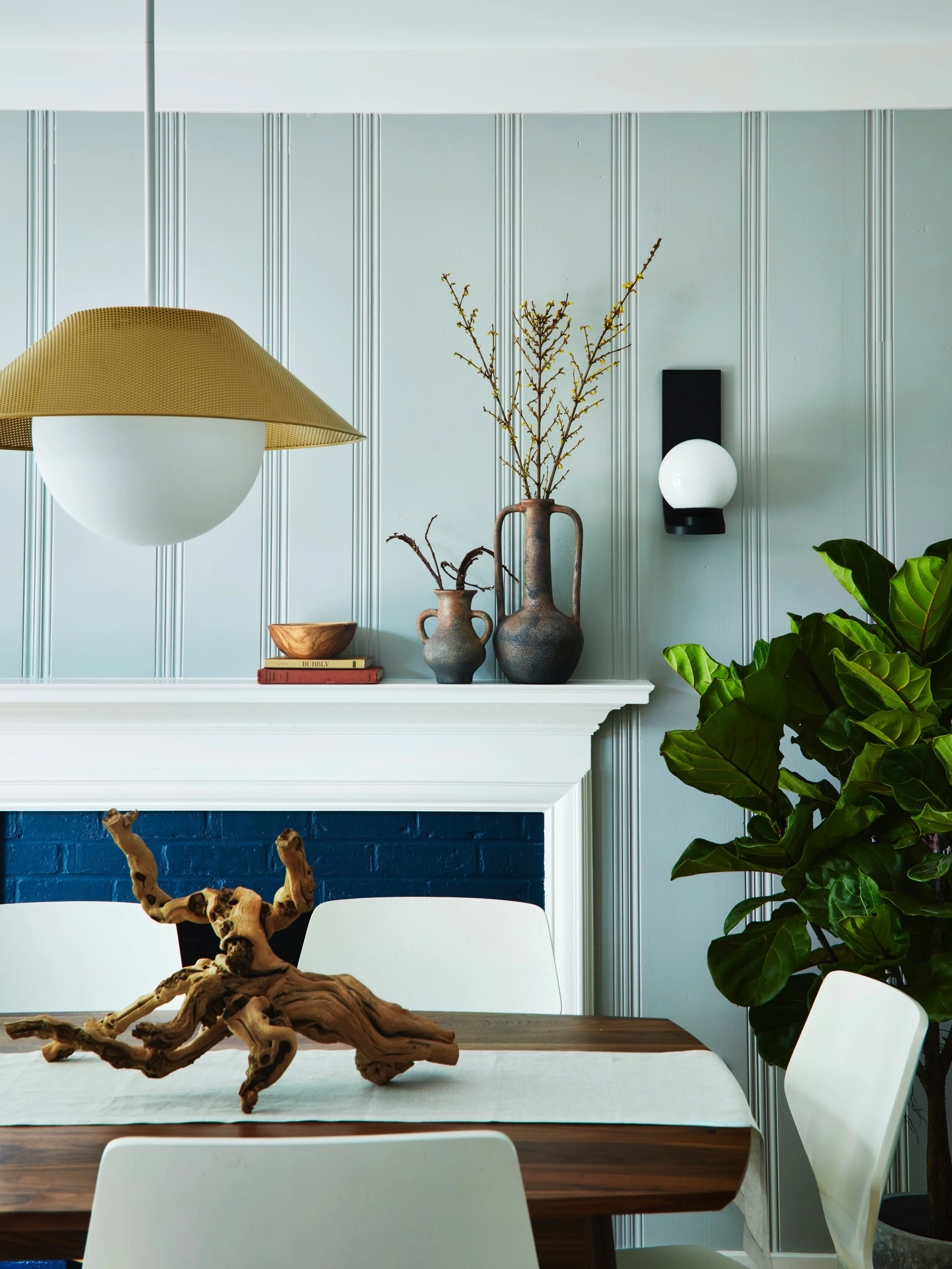 Modern dining room with a wooden table, white chairs, a large green plant, and decorative vases with branches on a mantle. A brass and white pendant lamp hangs above the table, and a wall-mounted light fixture with a spherical bulb is on the wall.