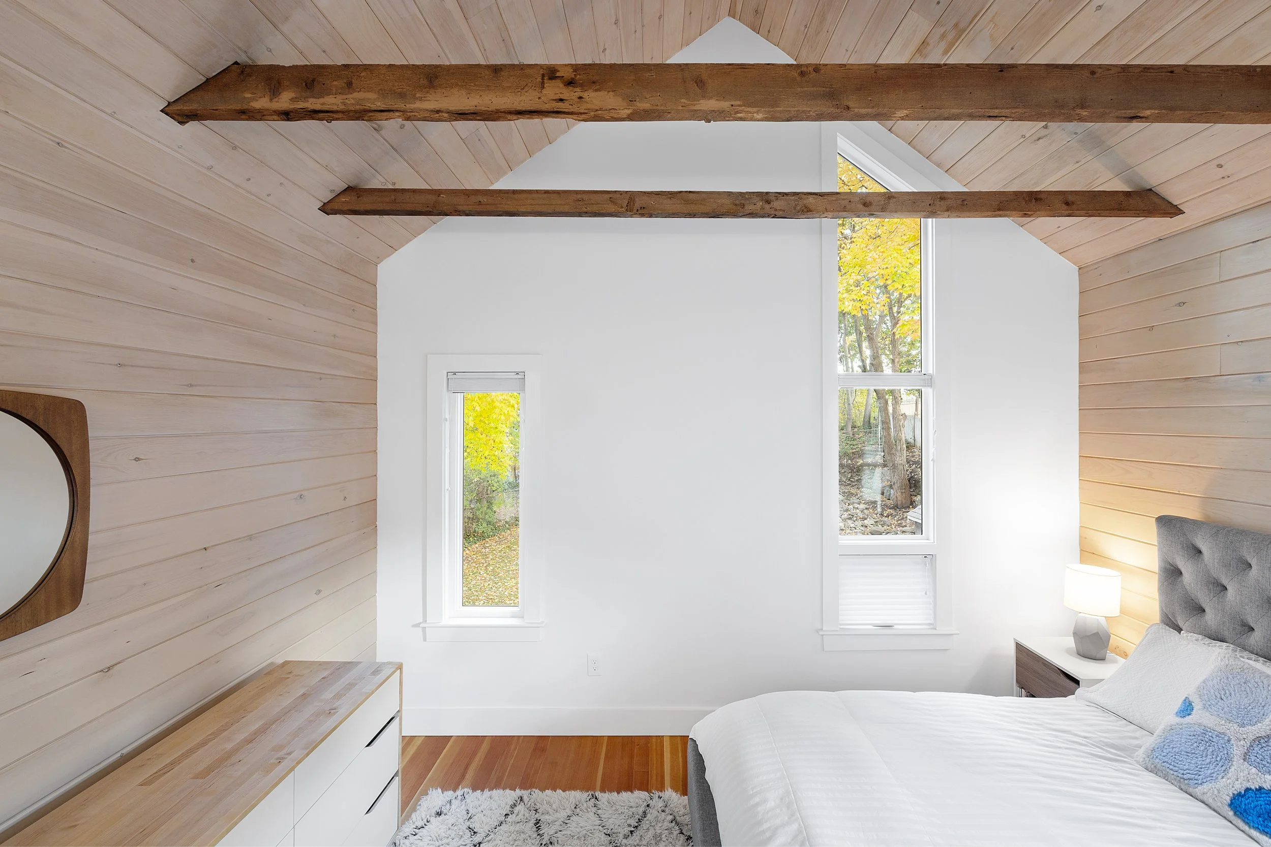 Bedroom with wooden ceiling beams, white wall, two tall windows with views of trees, bed with white bedding, nightstand with a lamp, and a gray headboard.