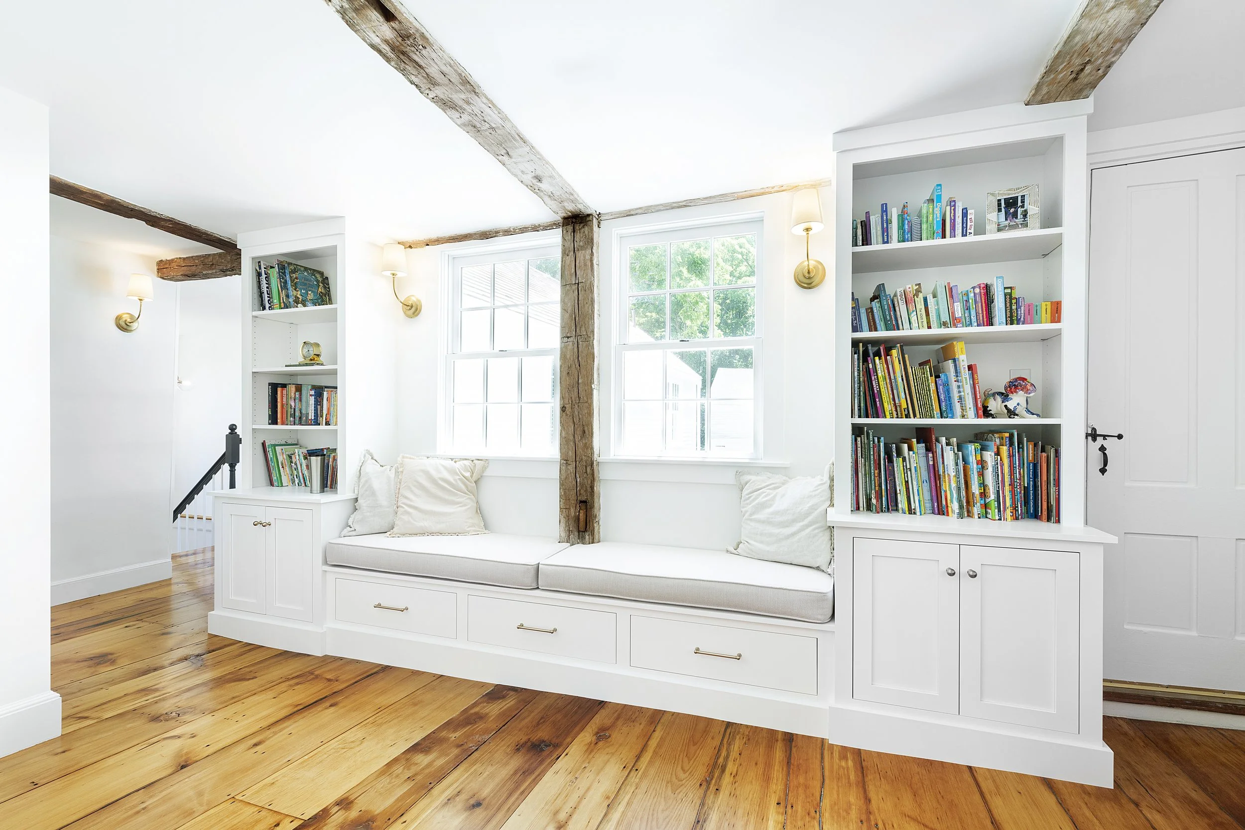 Bright reading nook with white built-in bench, pillows, two white bookshelves filled with colorful books, large windows, and wooden beams on the ceiling.