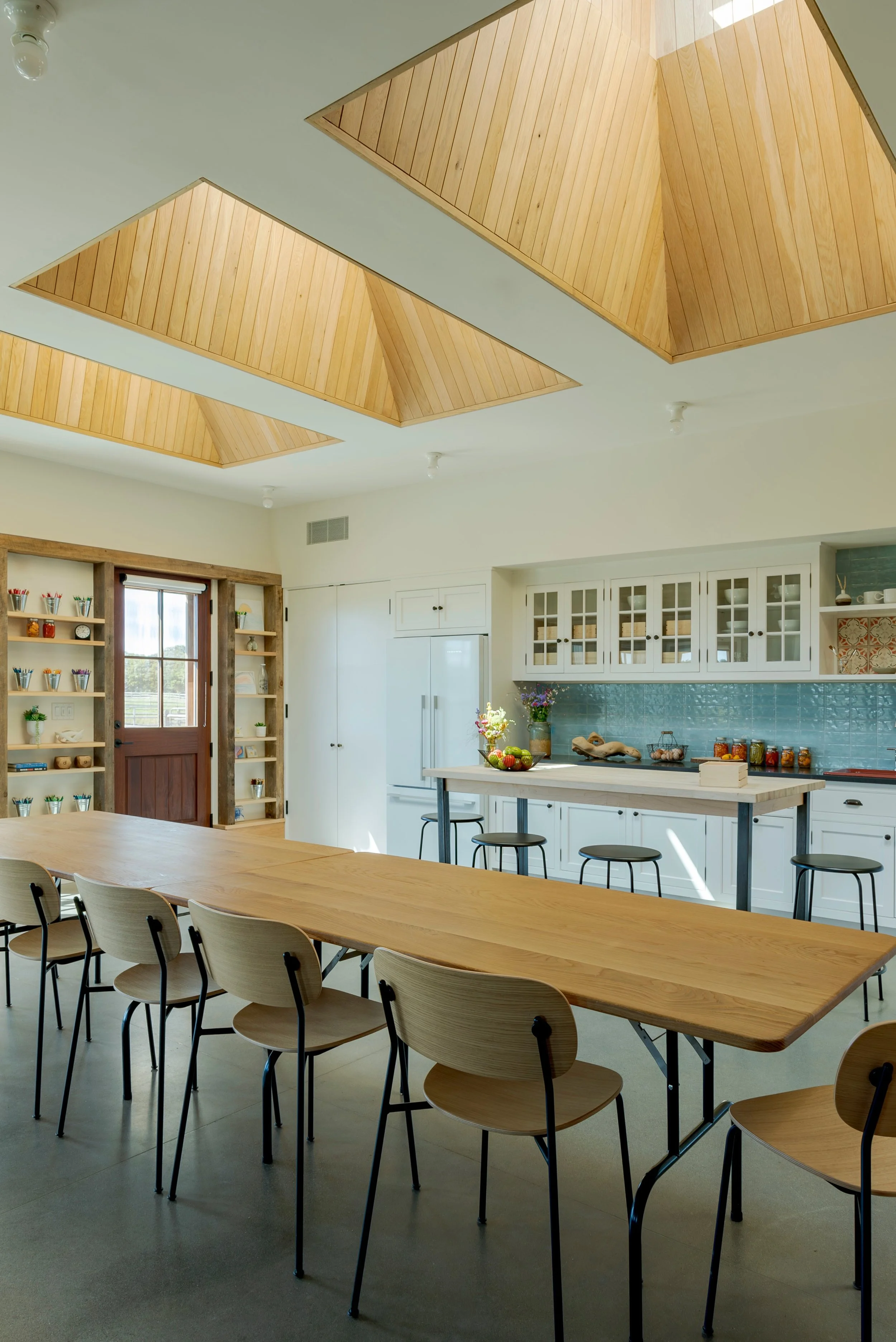 Interior view of a modern, minimalistic kitchen with a large wooden dining table, light wood chairs, white cabinetry, and unique wooden ceiling panels.