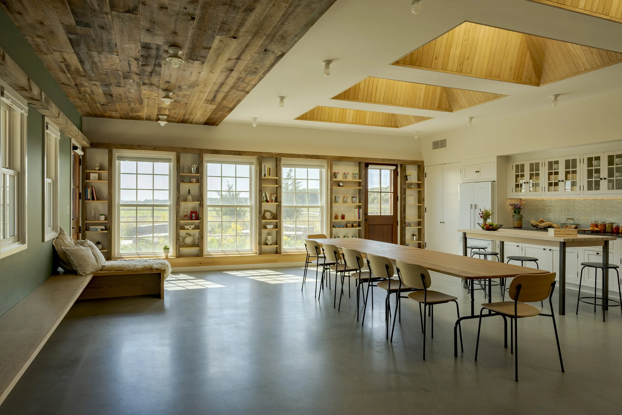 Bright, spacious kitchen and dining area with large windows, wooden ceiling accents, white cabinets, and a long wooden table with chairs.