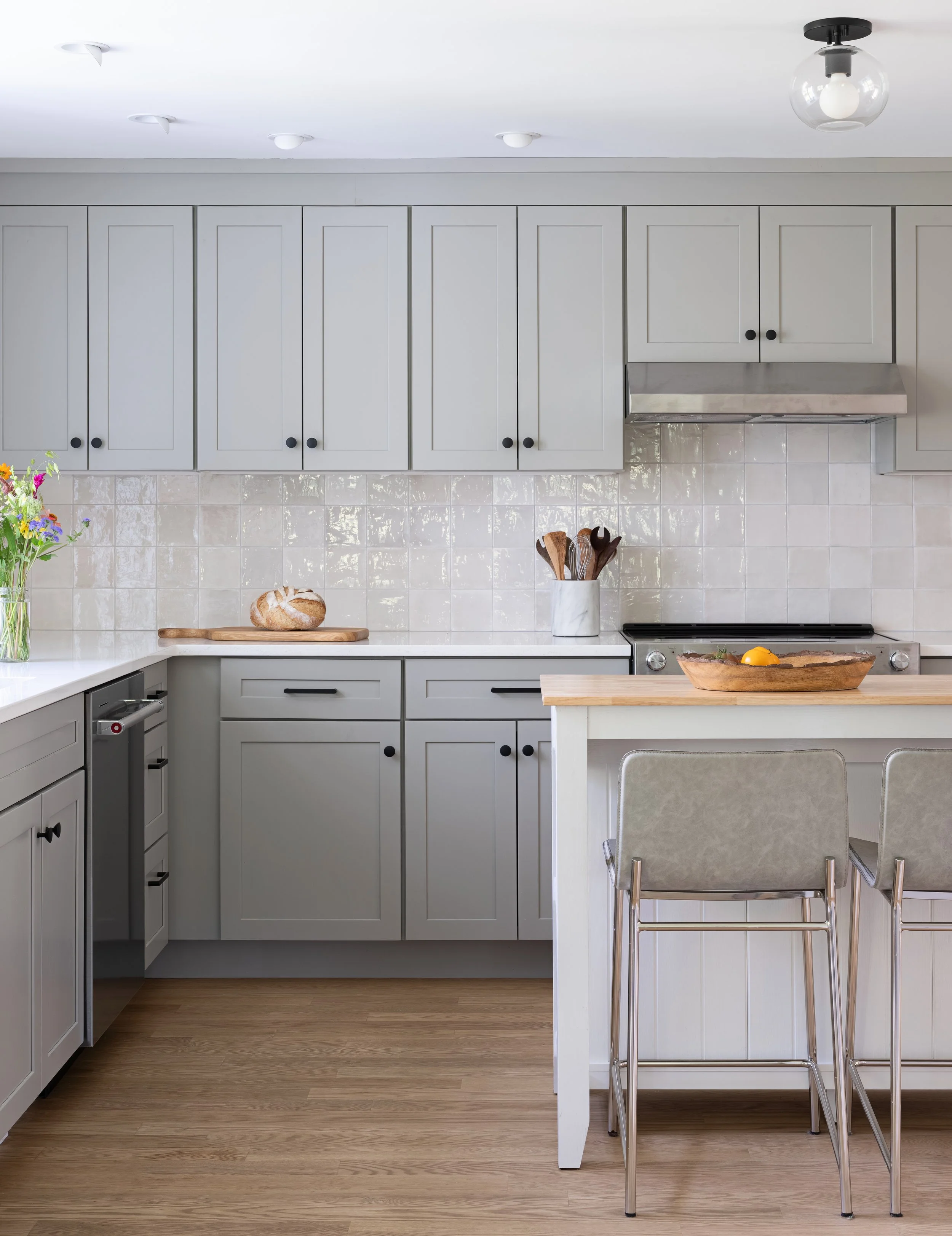 Modern kitchen with white cabinetry, a countertop with bread, a flower vase, kitchen utensils in a white marble container, a stovetop with an egg in a wooden bowl, and a breakfast bar with two chairs.