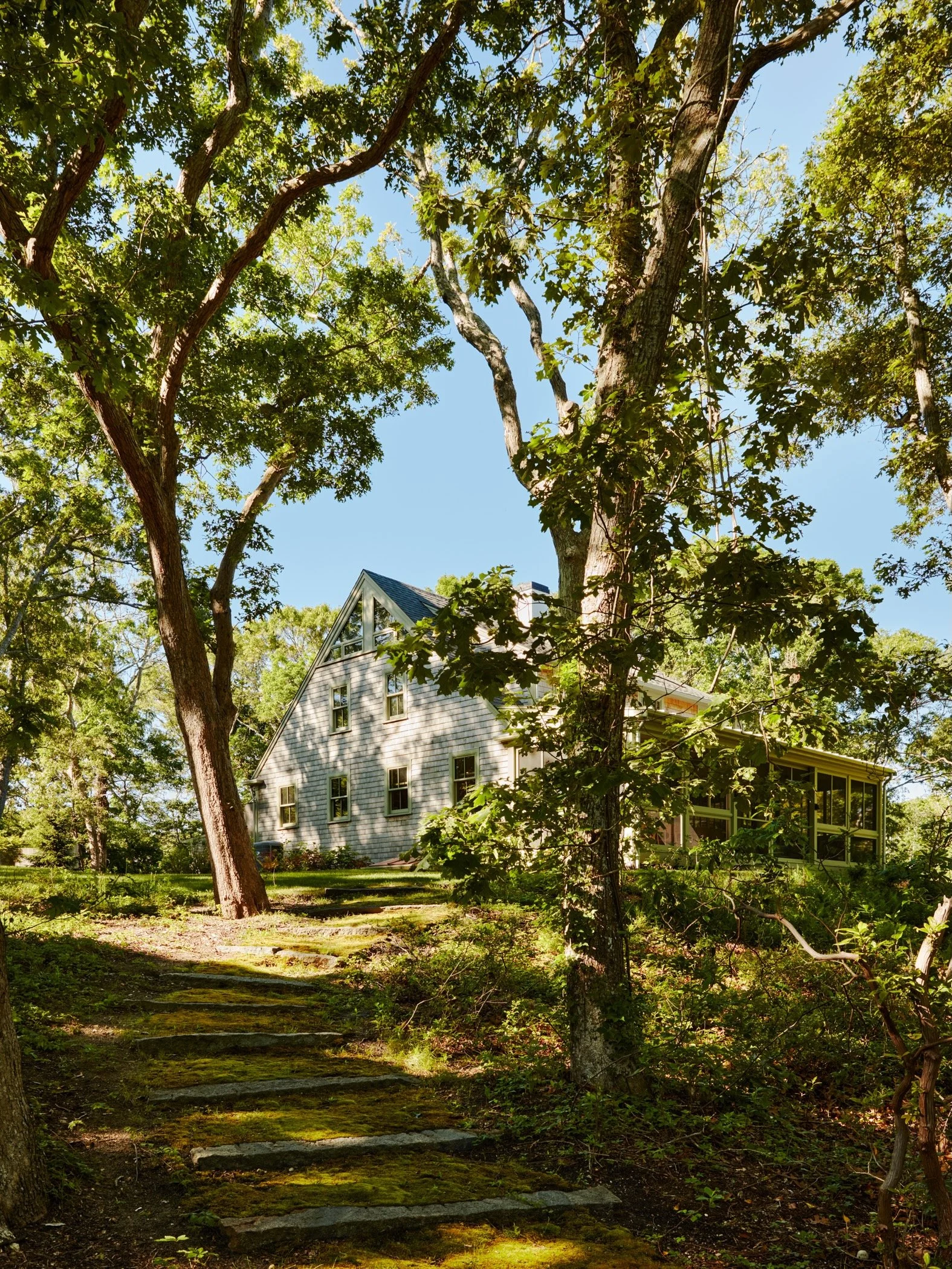 A house surrounded by trees on a sloped landscape with a stone staircase leading up to it, under a clear blue sky.