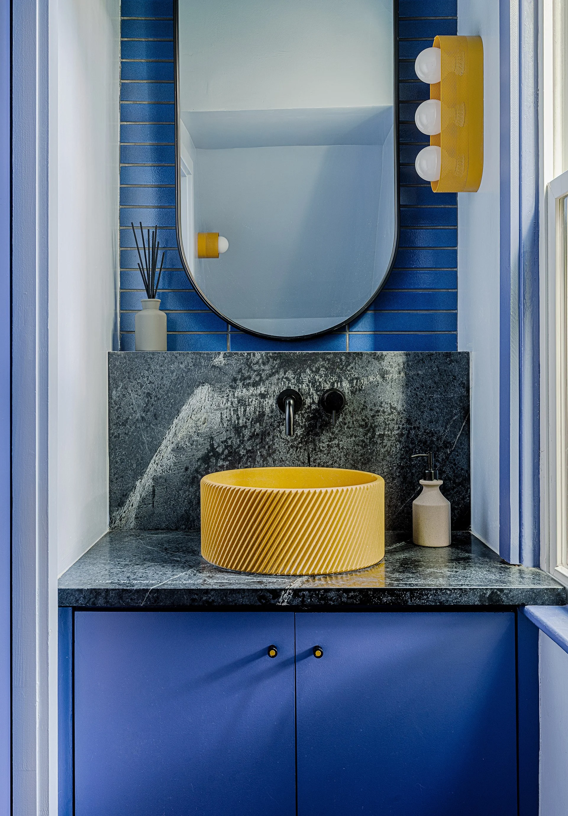 A modern bathroom vanity with a yellow textured sink, dark marble countertop, blue cabinets, a wall mirror, and a yellow light fixture with three bulbs. There is a white soap dispenser and a small white vase with black sticks. The wall behind has blu
