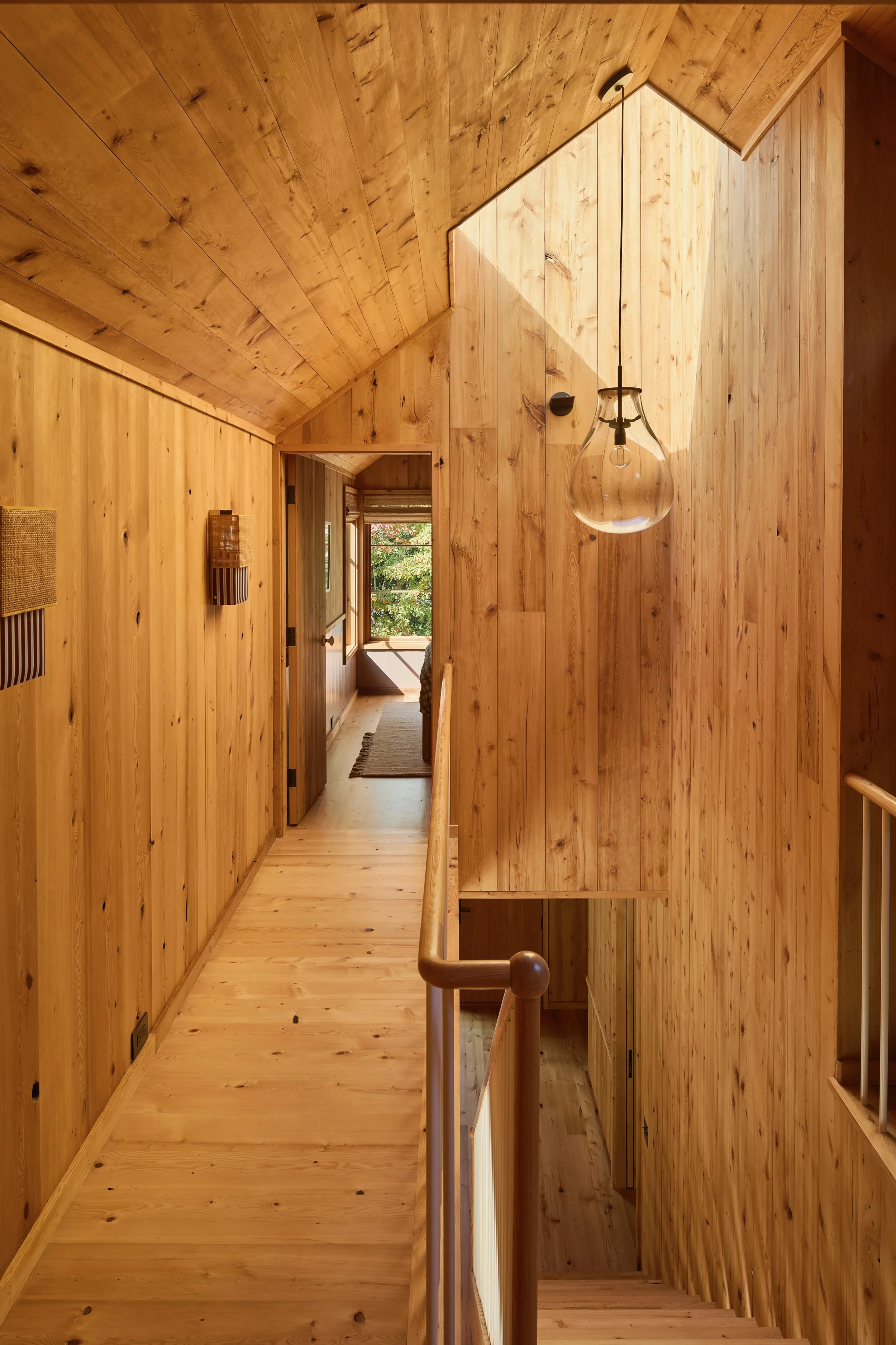 Wood-paneled hallway with staircase, natural light from a skylight and window at the end, and minimalist wall decor.