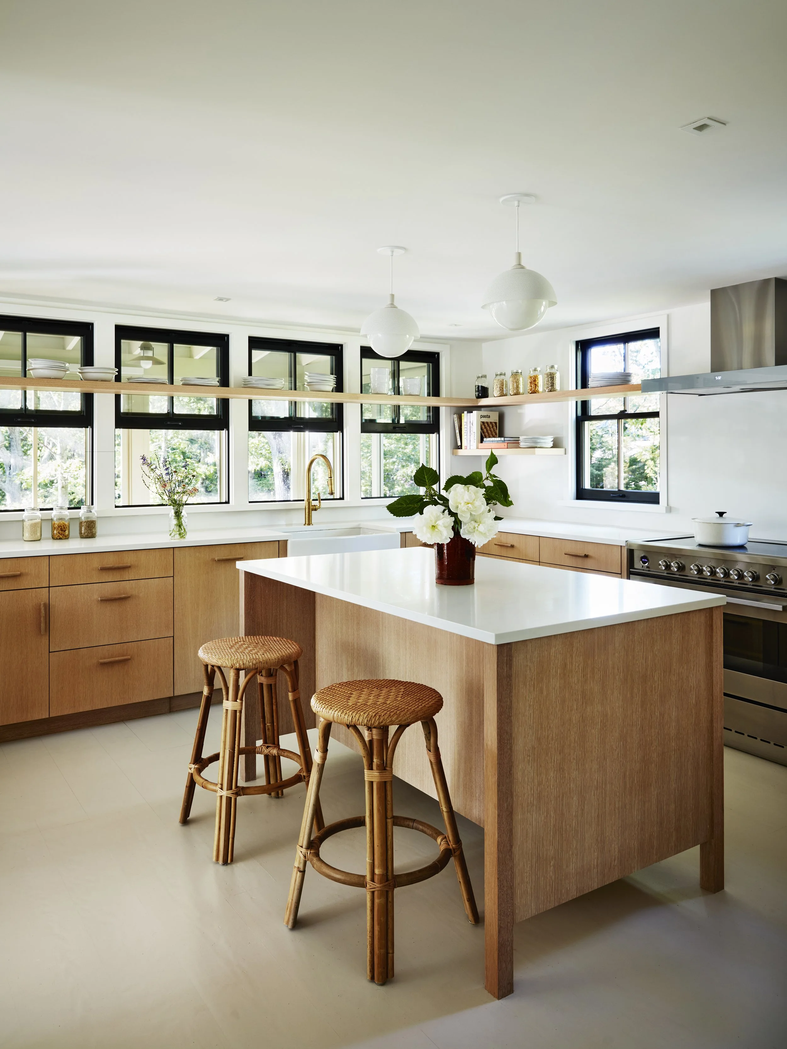 Modern kitchen with wooden cabinets, white island, and black-framed windows, decorated with a vase of white flowers and jars on the counter.
