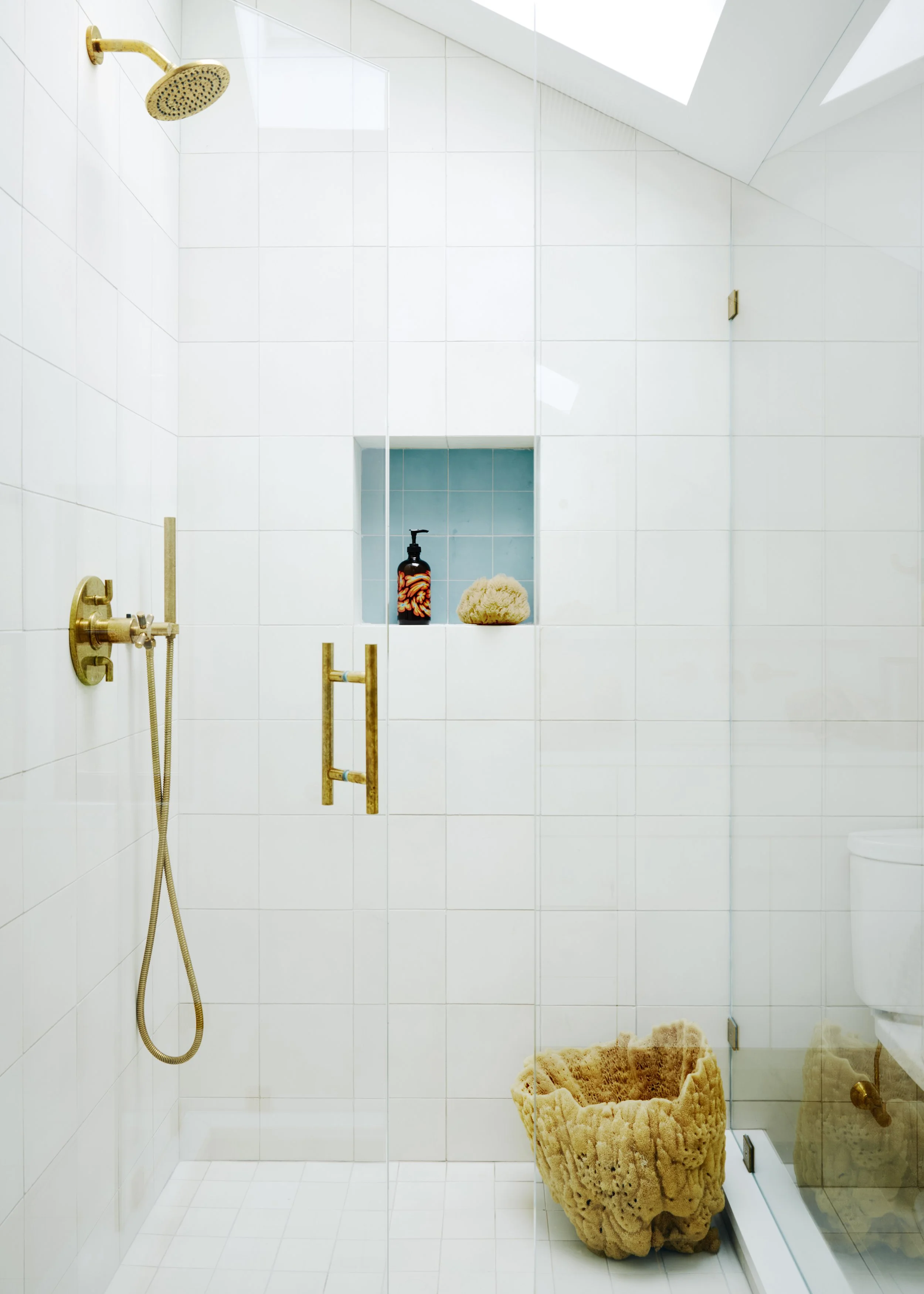 Modern white-tiled bathroom shower with gold fixtures including a rain shower head, a handheld shower, and a gold towel bar. A recessed shelf contains a soap dispenser and a loofah. A textured beige bath sponge rests on the shower floor, next to a gl