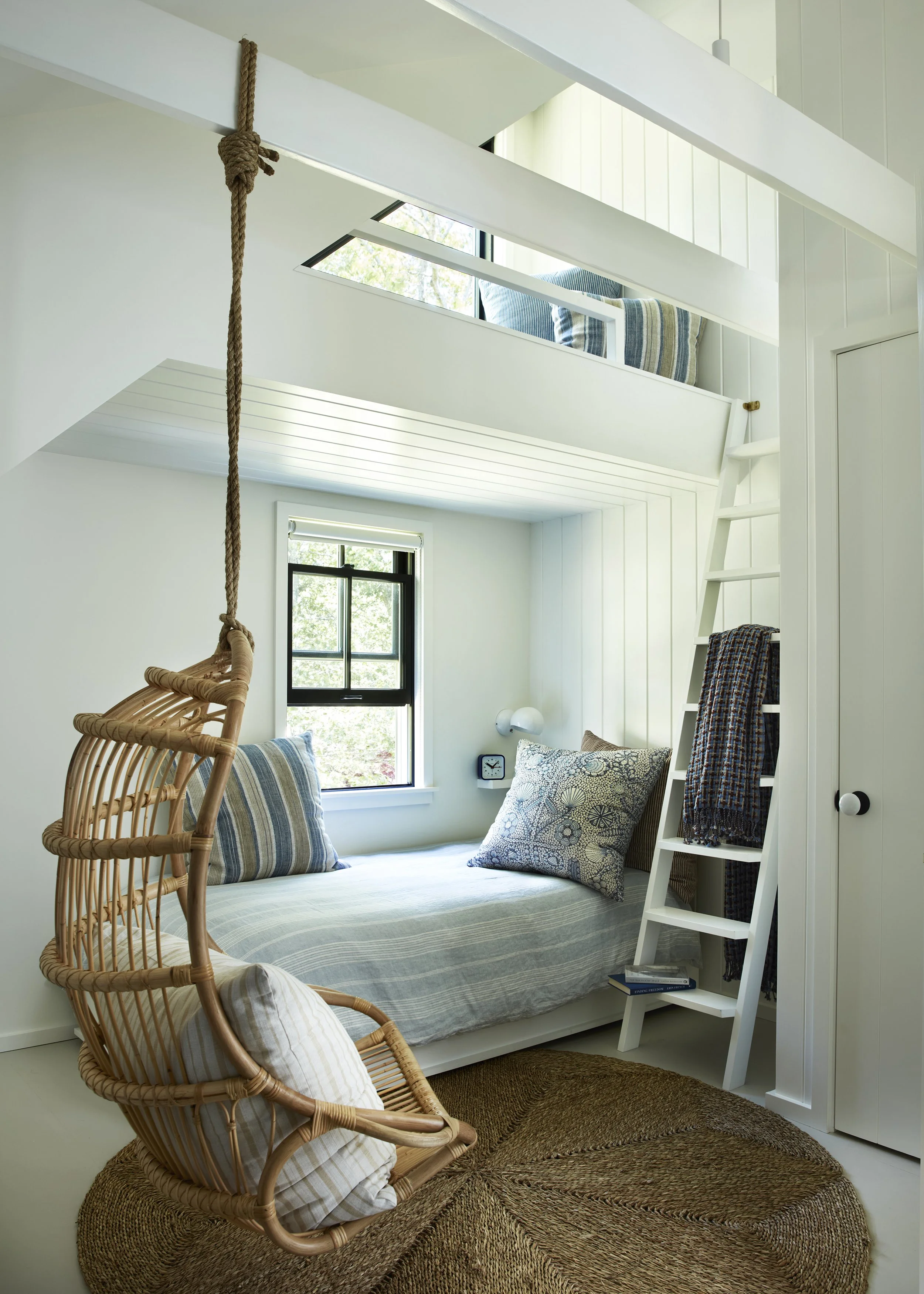 A cozy white bedroom with a built-in bed below a loft area. The lower bed has a striped bedspread and pillows, and a rattan hanging chair with a cushion is in front. A small window lets in natural light. The loft area has cushions and a blanket, with