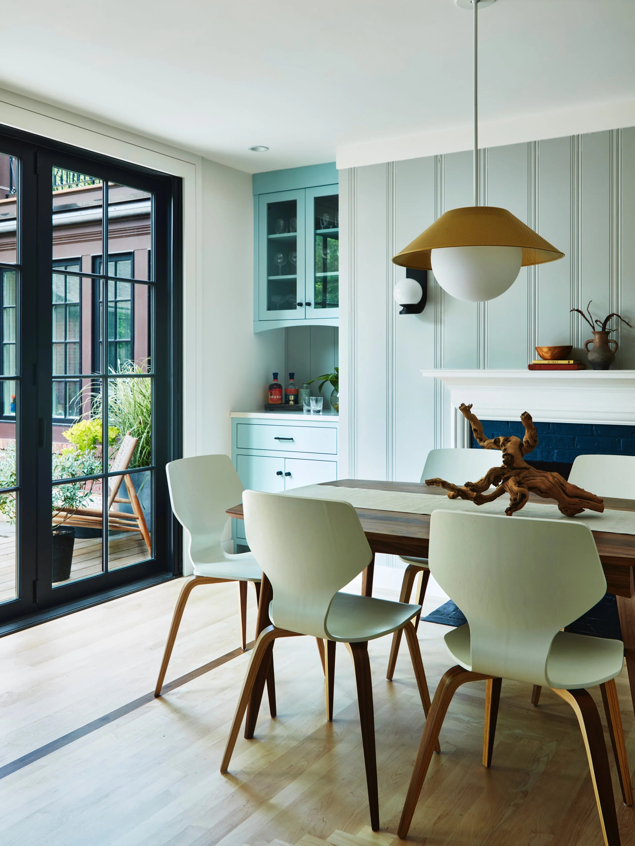 Modern dining room with a wooden table, white chairs, a hanging globe light with a brown shade, a blue cabinet, and large glass sliding doors leading to a patio with plants.