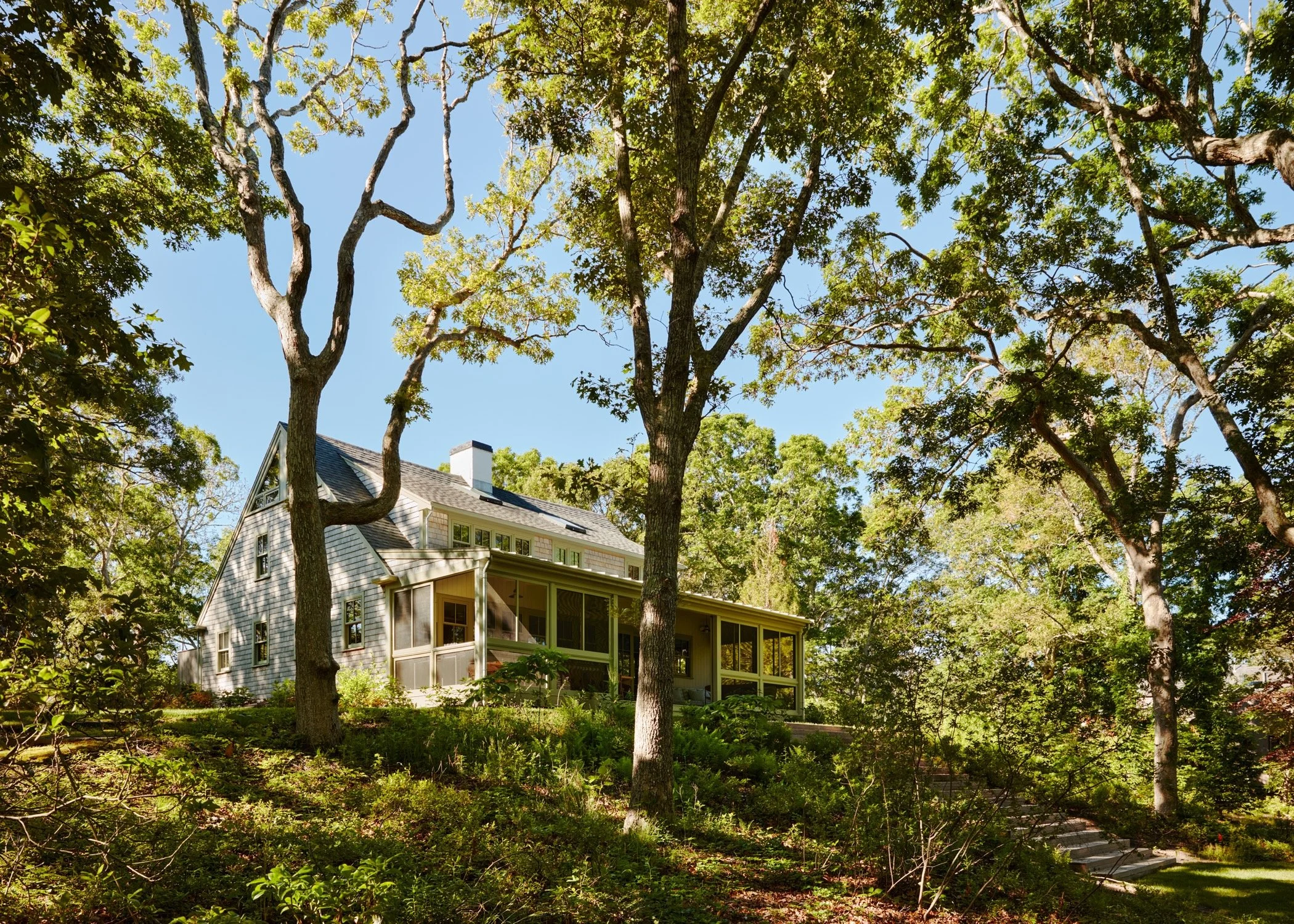 A two-story house with a screened porch nestled among tall trees on a lush green hillside.