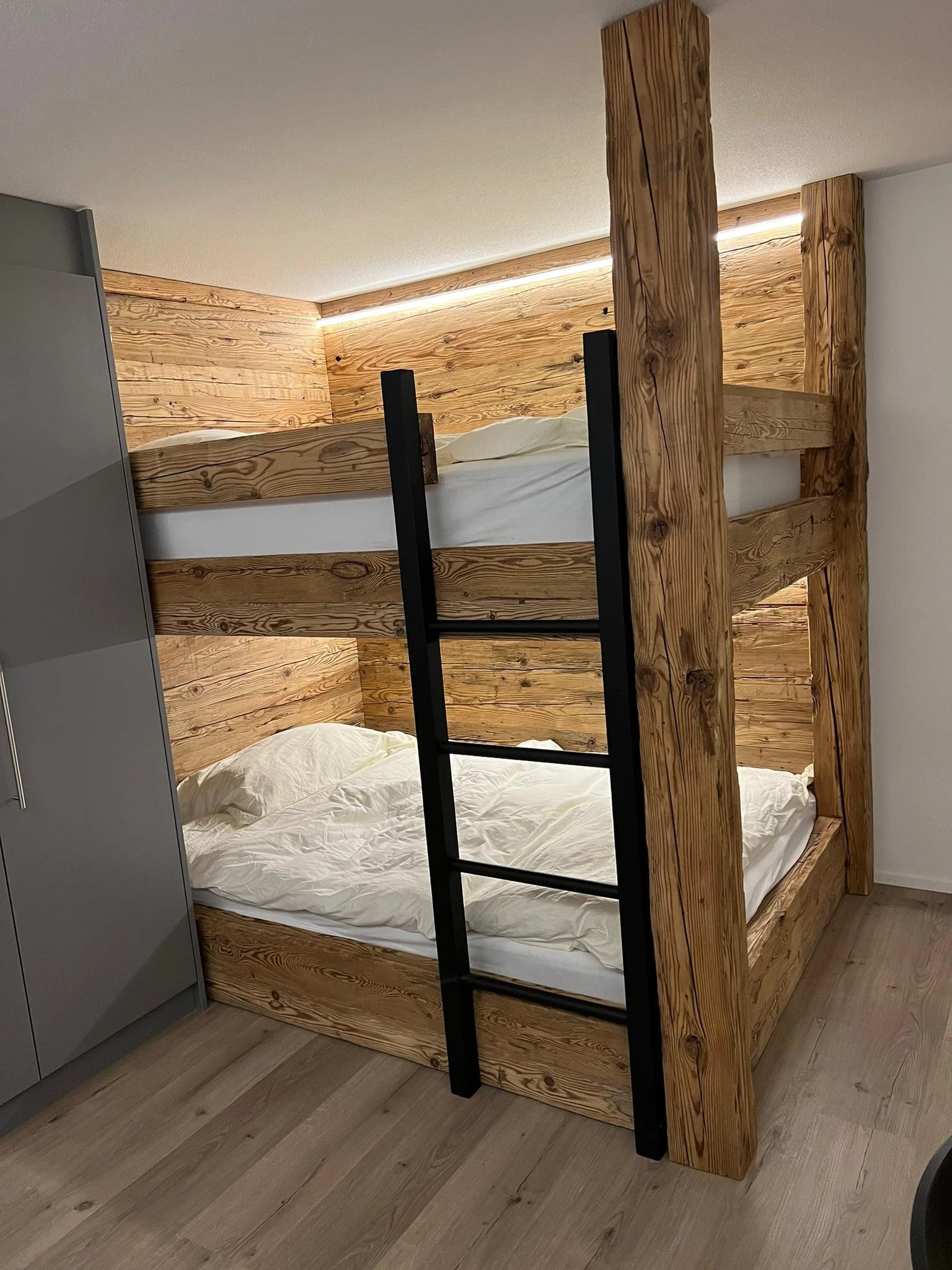 Ein Holzhochbett mit Leiter in einem modernen Schlafzimmer, braune Holzverkleidung auf der Wand im Hintergrund, neben einem grauen Schrank, auf Holzboden.