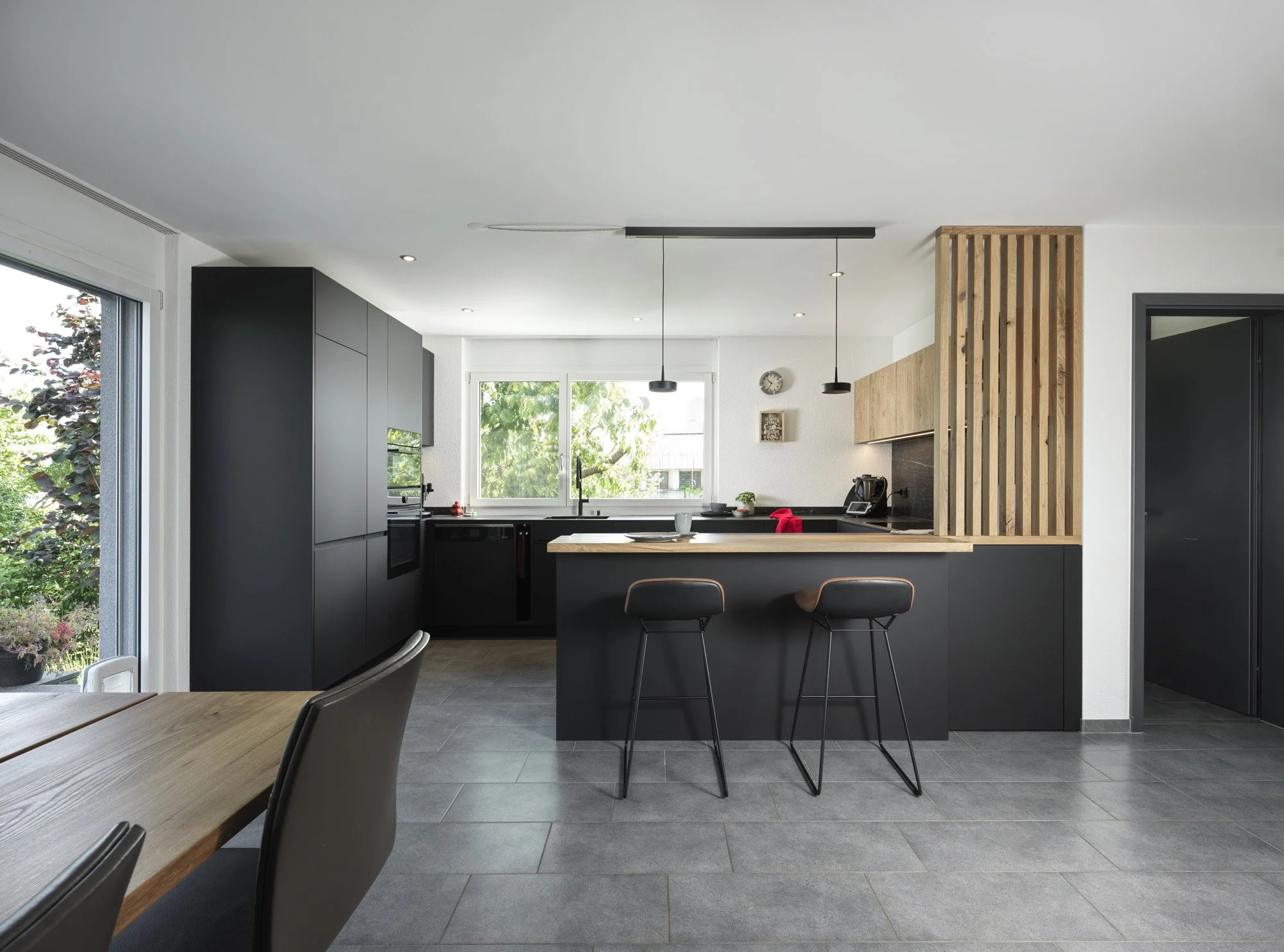Moderne schwarze und Holz-Küche mit Insel und Barhockern, Blick auf Fenster mit grüner Vegetation, minimalistische Gestaltung, helle Wände, dunkle Fliesenboden.