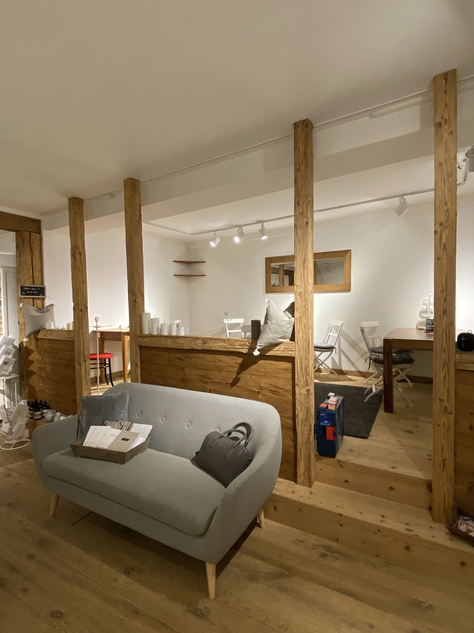 Innenraum eines Hauses mit Holzwänden und Möbeln, einer Couch mit Tasche und Zeitschriften, mehreren Stühlen, einem Schreibtisch, Dekorationsstücken und einer kleinen Treppe.