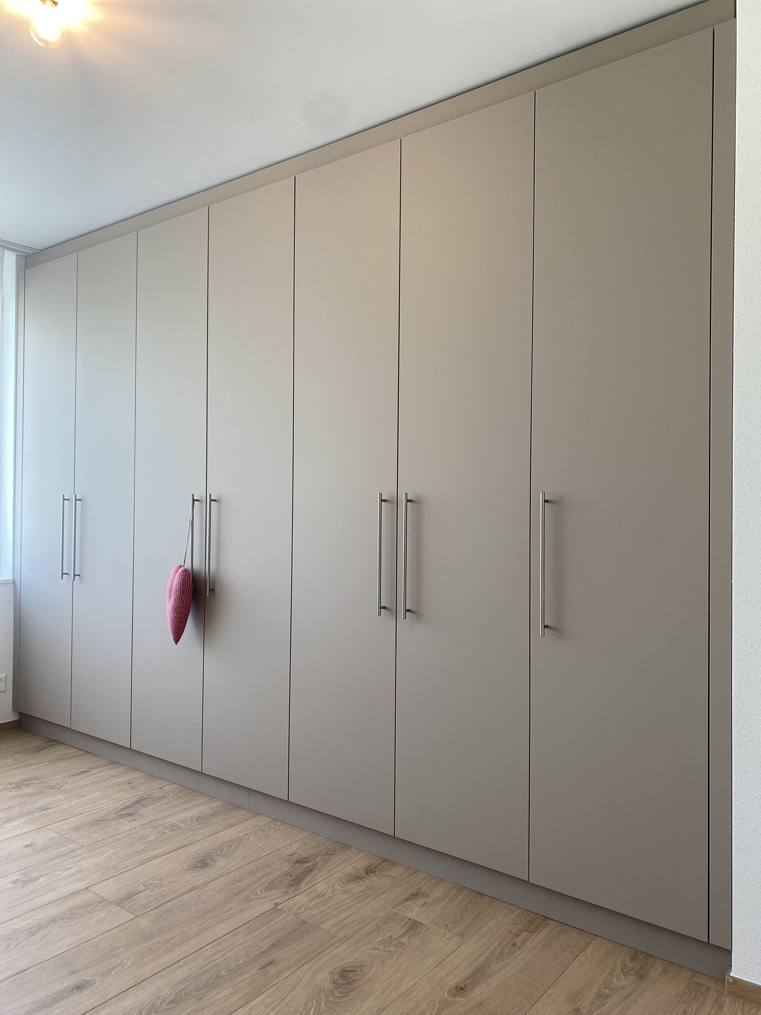 Großer, moderner Kleiderschrank mit mehreren weißen Türen und langen Metallgriffen in einem Raum mit Holzfußboden.