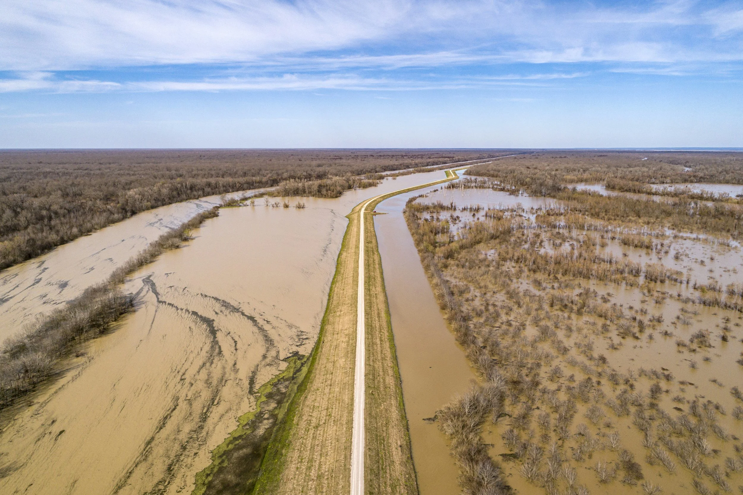 An aerial view of a flooded area with a straight road running through the middle, surrounded by water and bare trees under a partly cloudy sky.