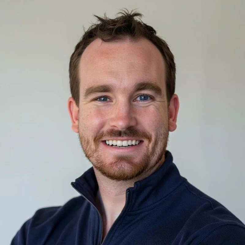 Headshot of a smiling man with brown hair and beard, wearing a dark jacket against a plain background.