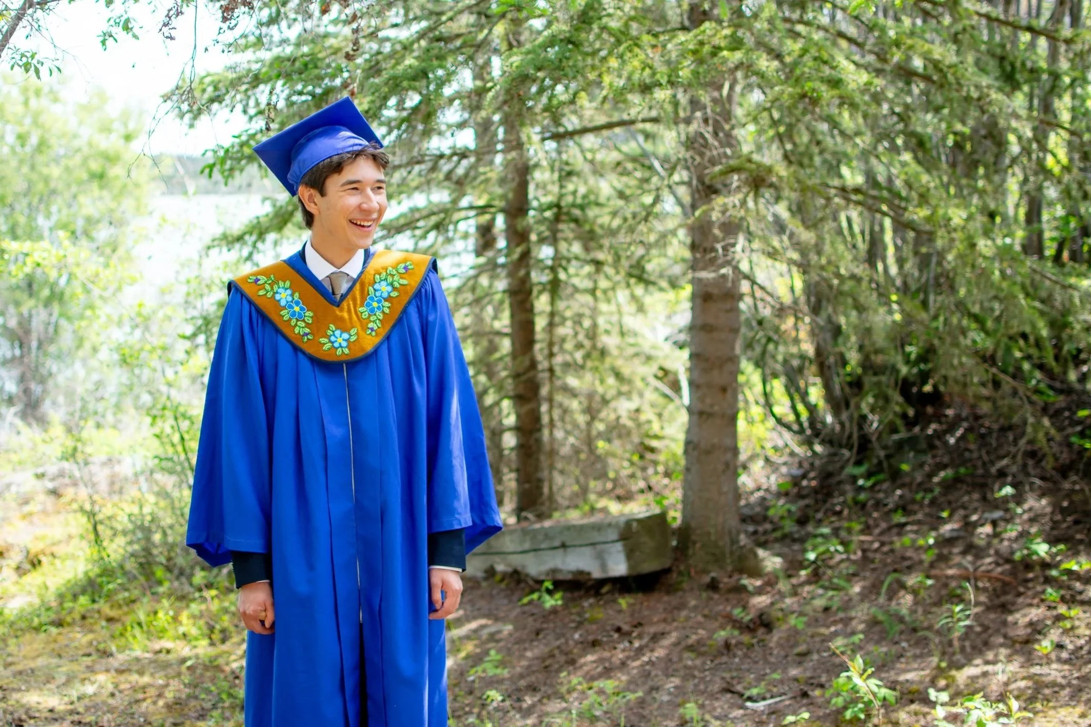 A young man in a blue graduation cap and gown, standing outdoors in a wooded area, smiling.