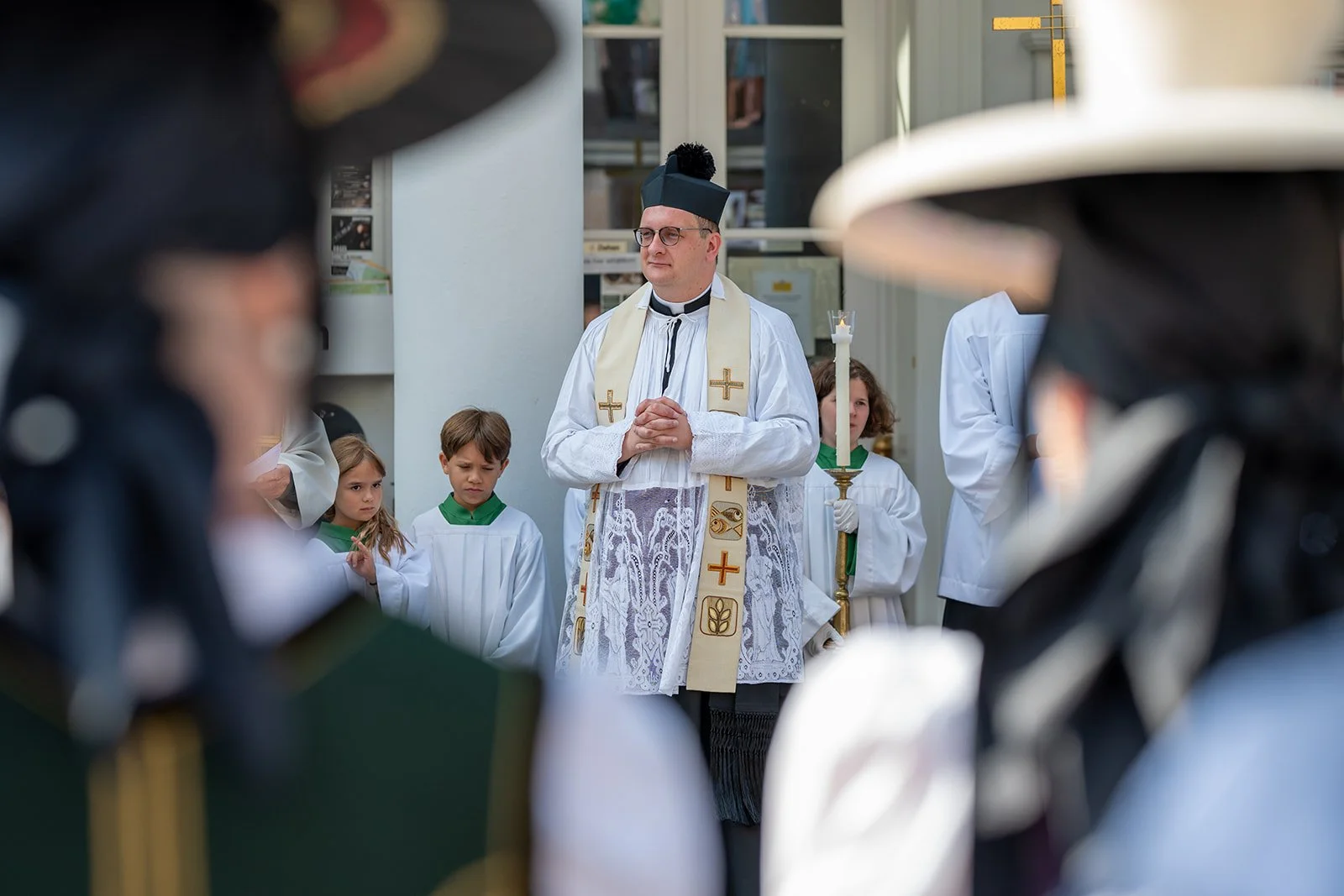 Ein Priester und Kinder bei einer religiösen Zeremonie, umgeben von Menschen in festlicher Kleidung, vor einem Gebäude.