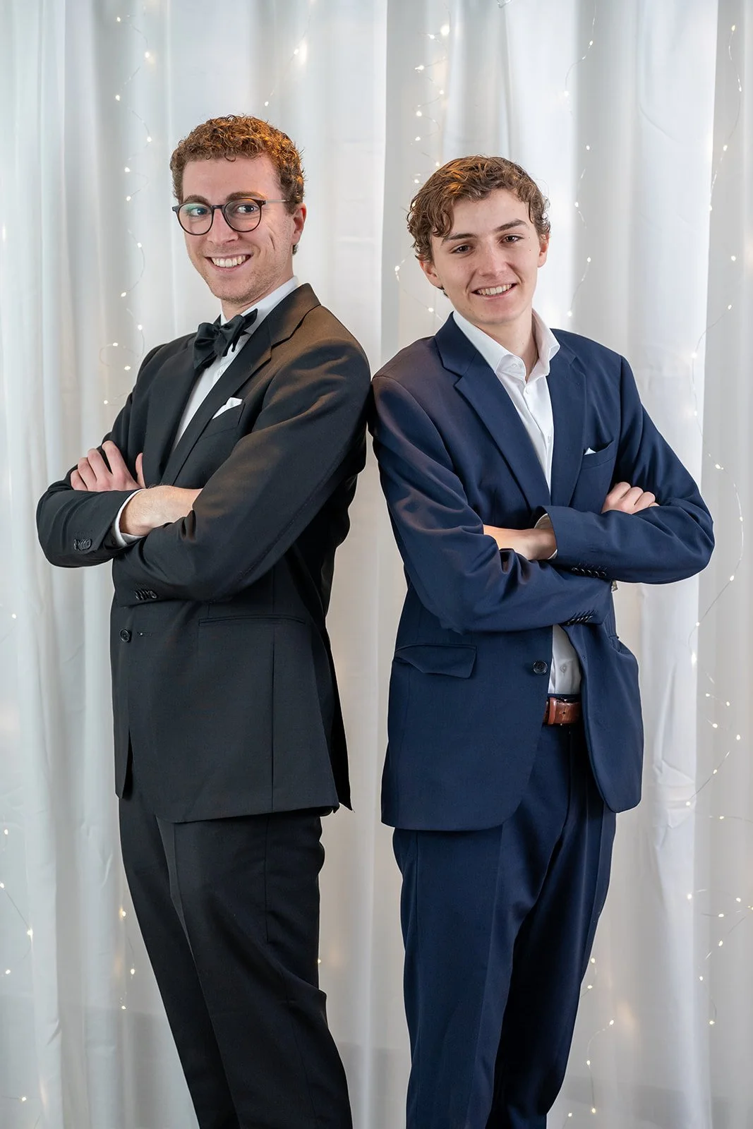 Zwei junge Männer in Anzügen stehen Rücken an Rücken vor einem weißen Vorhang mit Lichterdekoration, beide mit verschränkten Armen und lächeln.