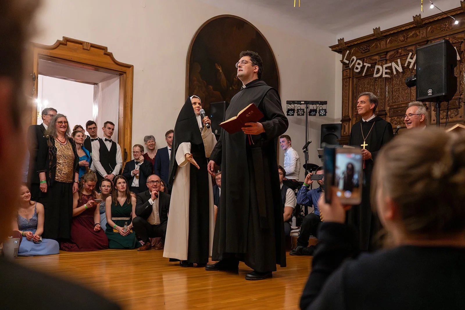 Ein religiöses Zeremonie mit einem Mönch, einer Nonne und einem Priester in einer Kirche, umgeben von Gästen, die das Ereignis beobachten und Fotos machen.