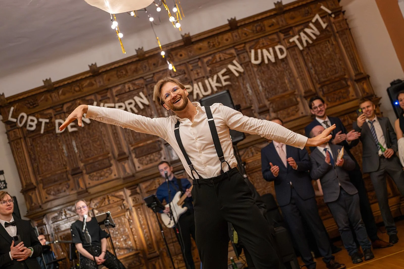 Ein Mann tanzt fröhlich bei einer Feier, im Hintergrund stehen mehrere Männer und Frauen in Anzügen und Kleidern, einige applaudieren, eine Person spielt Gitarre, die Raumdekoration ist Holzreich mit einer aufgehängten Banner mit deutschen Worten.