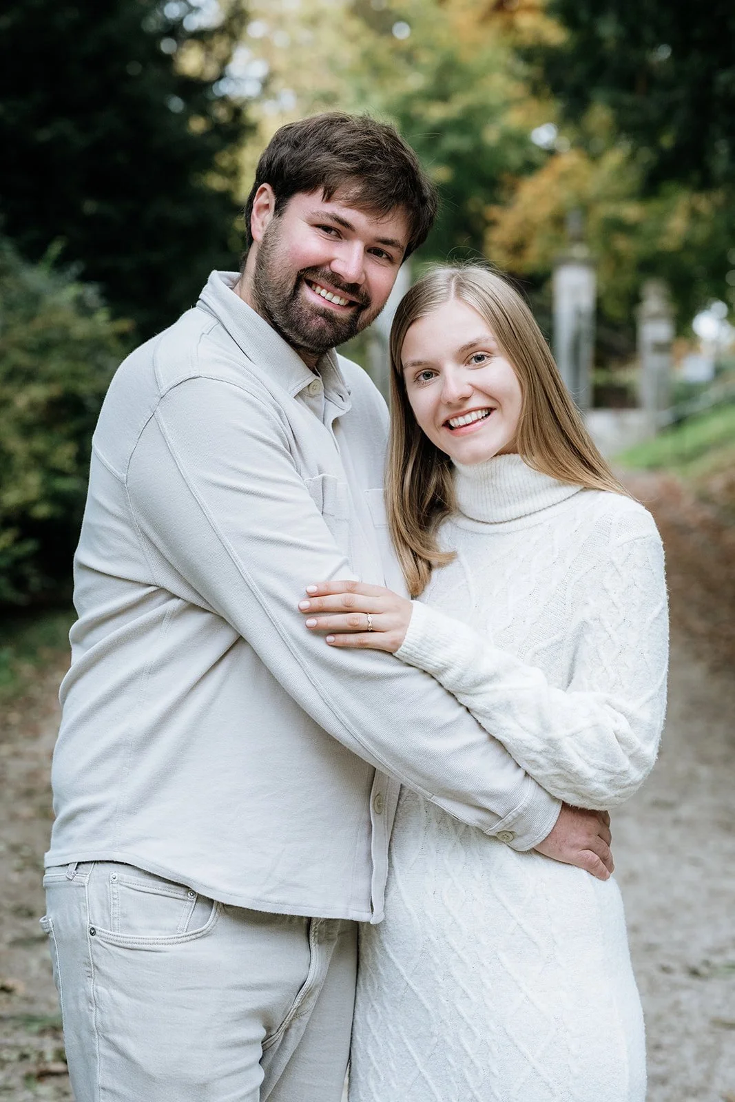Ein Paar, ein Mann und eine Frau, lächeln und umarmen sich im Freien in einer Parkumgebung mit grünen Bäumen im Hintergrund.