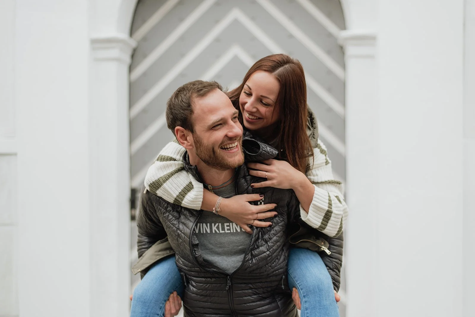 Ein lachendes Paar in legerer Kleidung, das sich gegenseitig trägt, vor einem modernen weißen Gebäude.