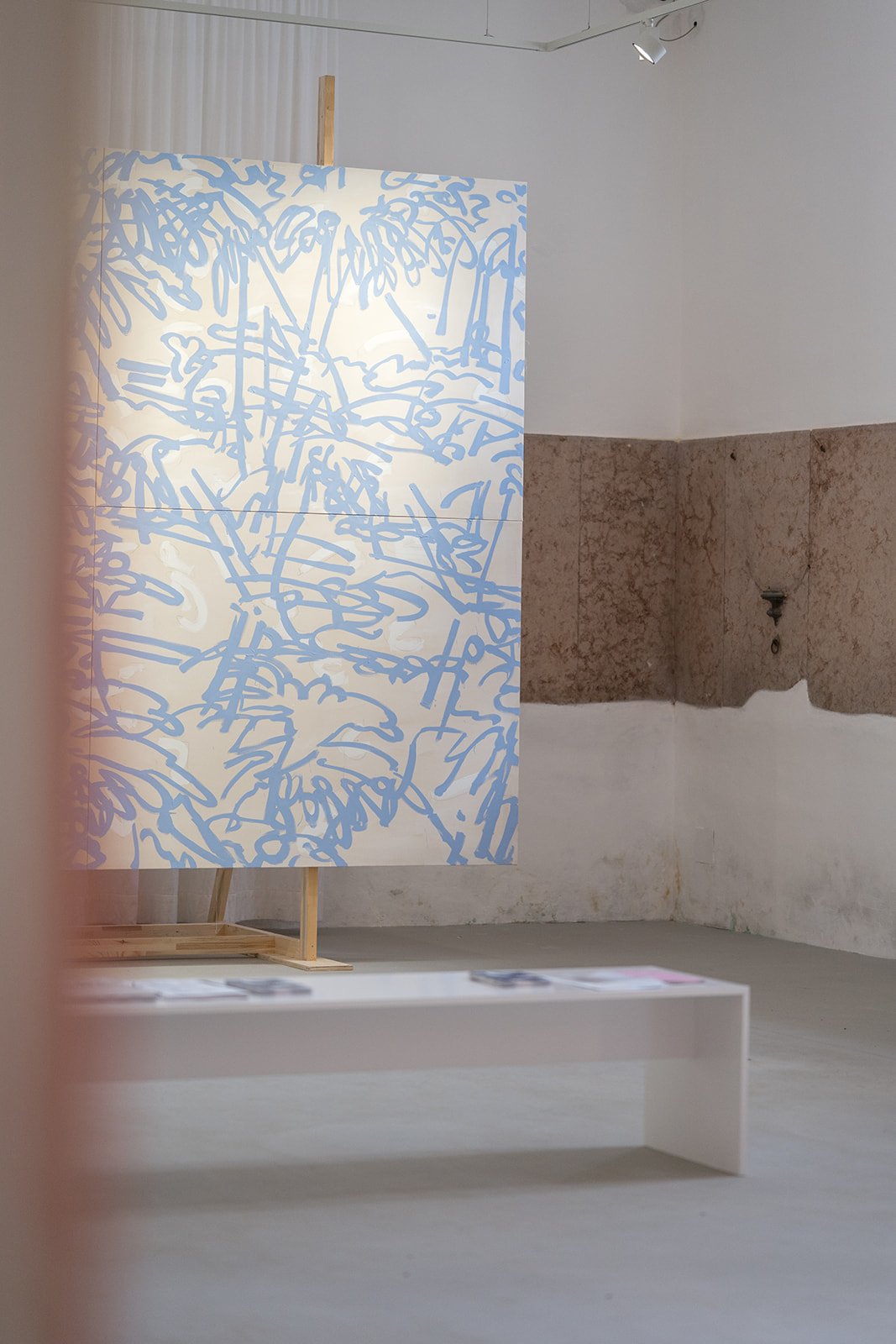 Eine Kunstinstallation mit einer großen, weißen Platte, die mit blauen, abstrakten Linien bemalt ist, steht in einem minimalistischen Raum mit weißen und braunen Wänden.