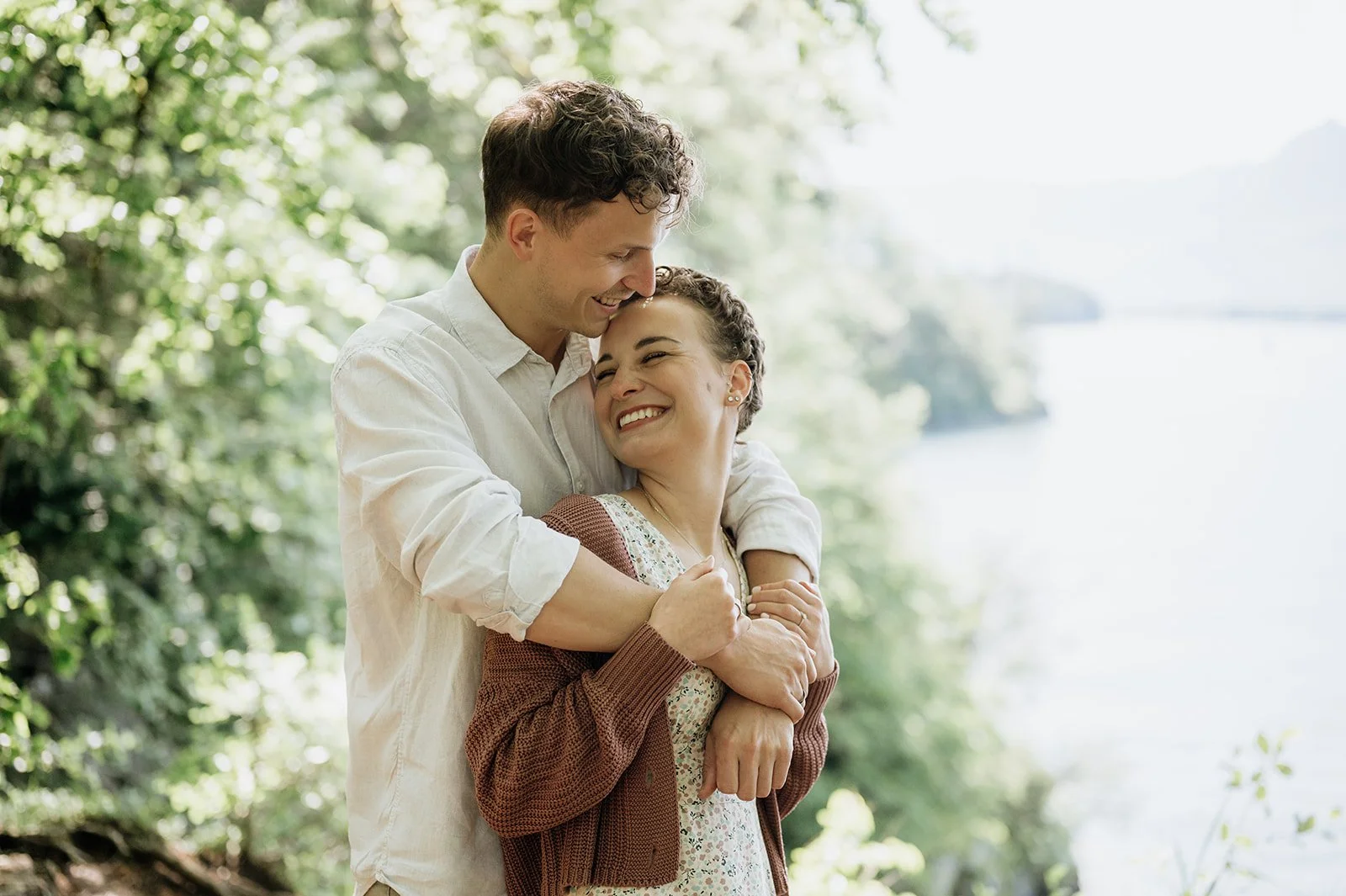 Paar umarmt sich lachend an einem sonnigen Tag im Grünen am Fluss.