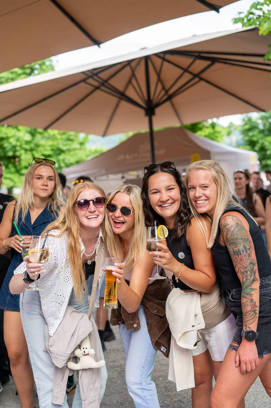 Gruppe junger Frauen, die bei einer Feier im Freien lachen, trinken und Spaß haben, mit großen Sonnenschirmen im Hintergrund.