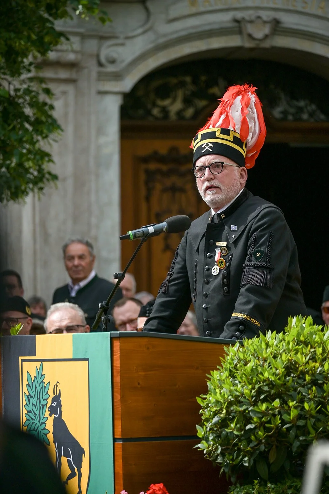 Ein älterer Mann mit Brille, in einer schwarzen Uniform mit Orden und einem schwarzen Hut mit rotem und weißen Federbusch, spricht an einem Rednerpult bei einer Zeremonie in einer historischen T-Kirche.