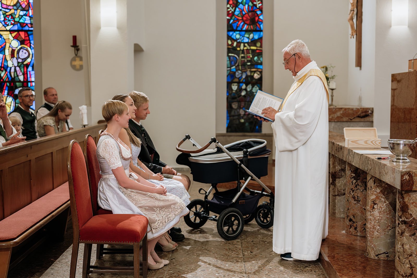 Ein Priester steht vor einer Gruppe von Kindern und Erwachsenen in einer Kirche während einer Taufe. Der Priester liest aus einem Buch, während die Kinder auf Sitzen vor ihm sitzen. Im Hintergrund sind bunte Kirchenfenster zu sehen.