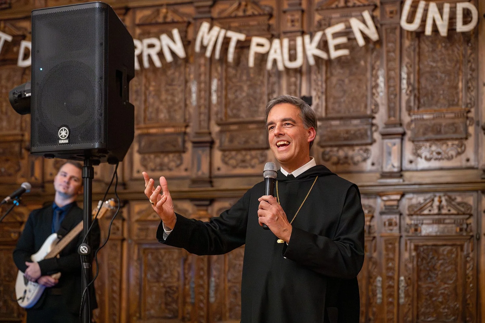 Mann in schwarzer Robe mit Mikrofon, lachend auf einer Bühne mit Holzverzierung, im Hintergrund Banner mit deutschem Text.