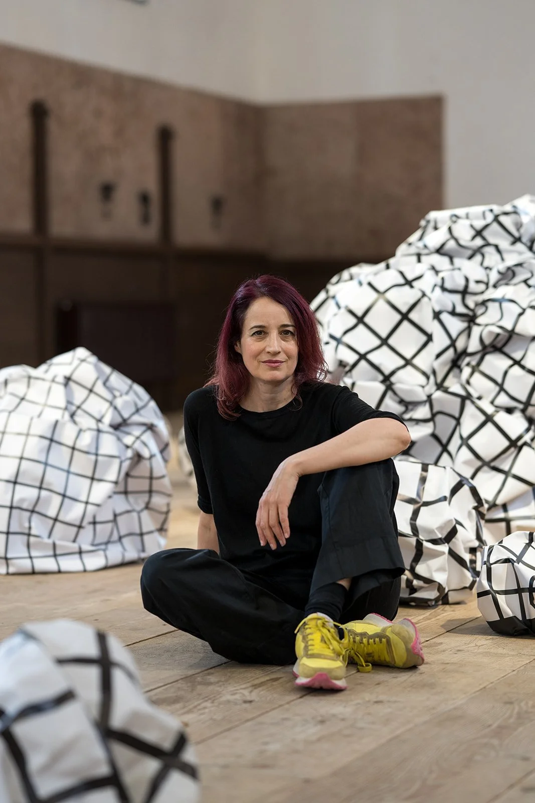 Eine Frau mit roten Haaren, schwarz gekleidet, sitzt auf einem Holzboden, umgeben von weißen, gecheckten Plastiktüten, und blickt in die Kamera.
