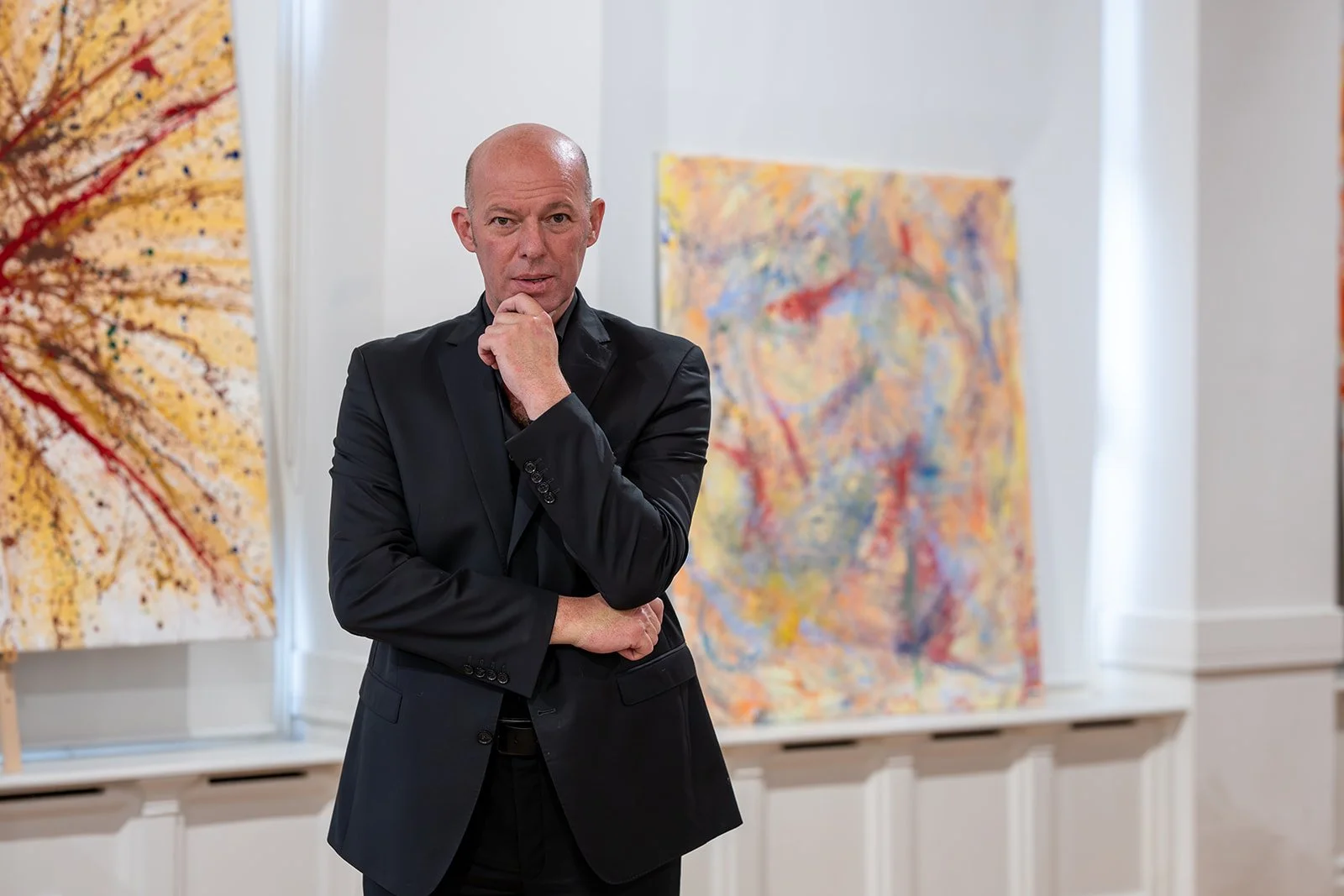 Ein Mann in schwarzer Anzug steht vor bunten abstrakten Gemälden in einer Kunstgalerie.