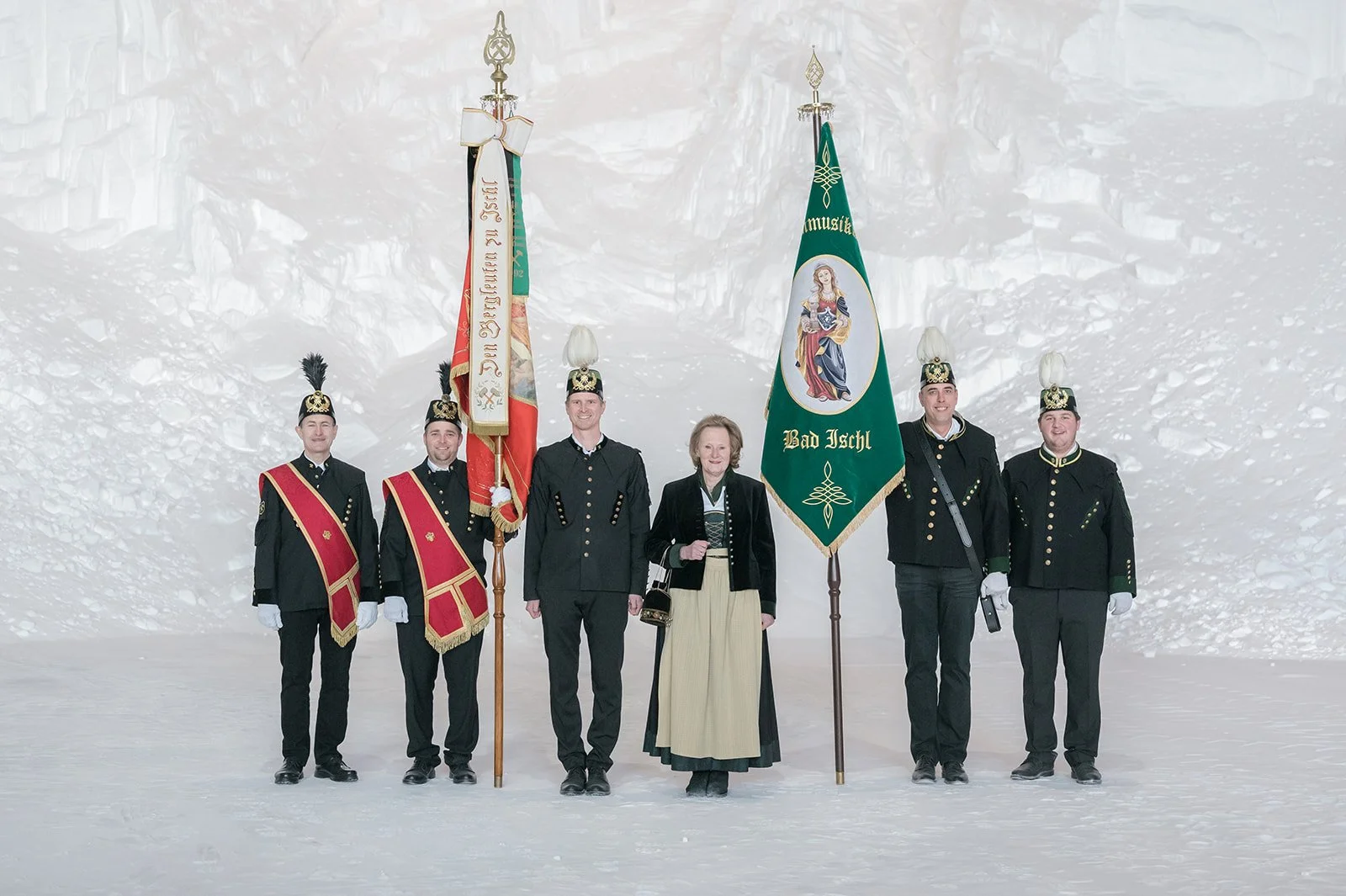 Fünf Personen in traditionellen Trachten und Uniformen stehen in einer Schneelandschaft, zwei halten Flaggen, die andere drei stehen aufrecht und lächeln.