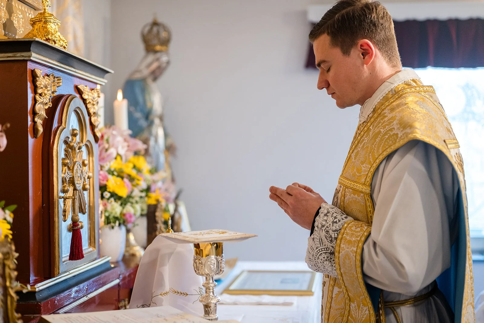 Ein Priester in goldener liturgischer Kleidung betet in einer Kirche vor einem Altar mit Blumen und religiösen Gegenständen.