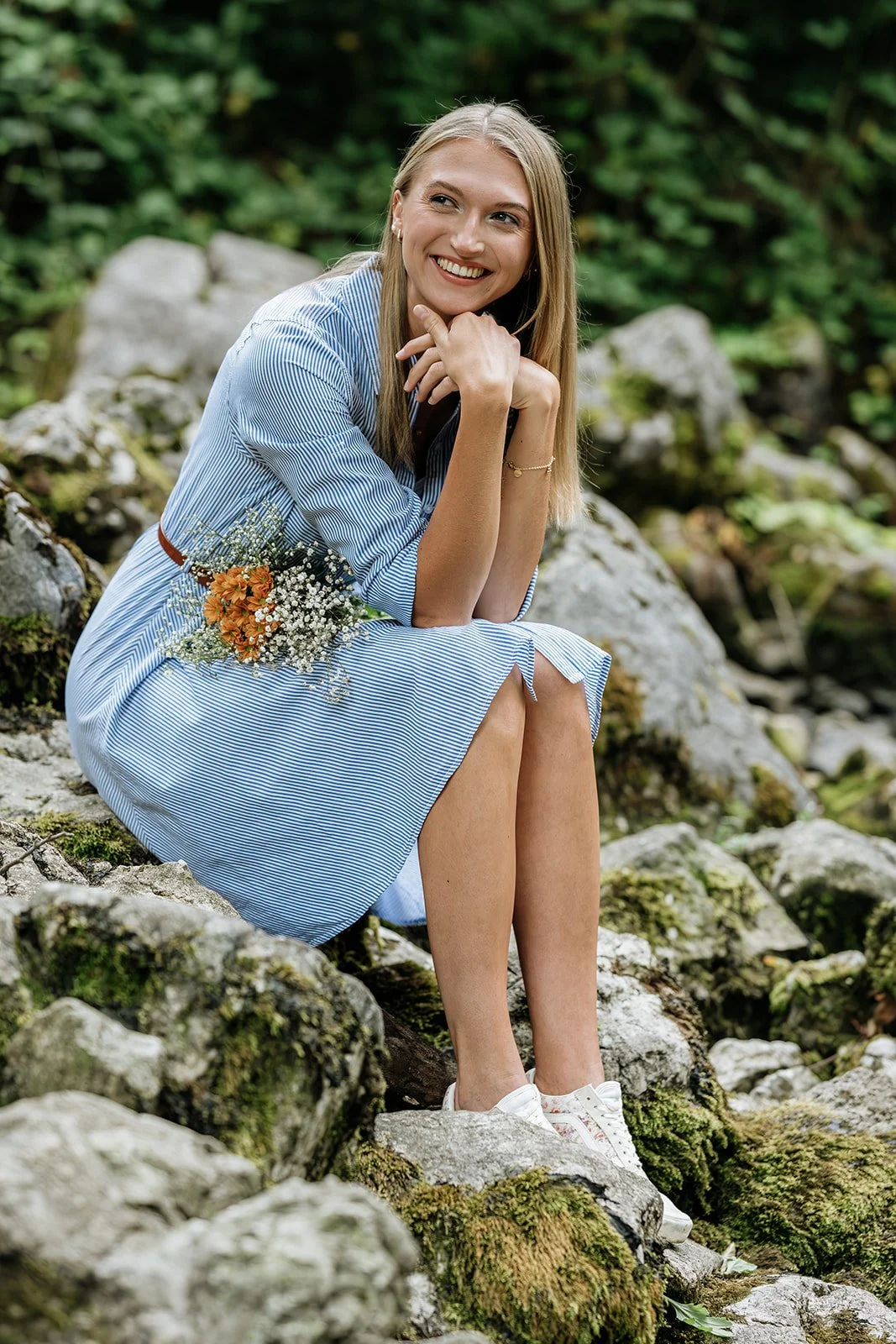 Lächelnde junge Frau in blau-weiß gestreiftem Kleid mit Blumen sitzt auf Steinen in der Natur.