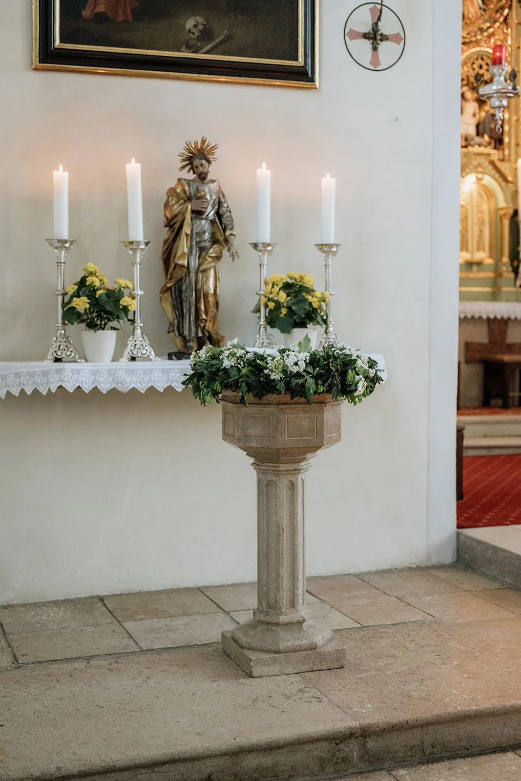 Ein Altar mit einer Jesus-Statue, vier weiße Kerzen, zwei Pflanzen mit gelben Blumen, und ein Blumenarrangement auf einem gotischen Steinsockel in einer Kirche.