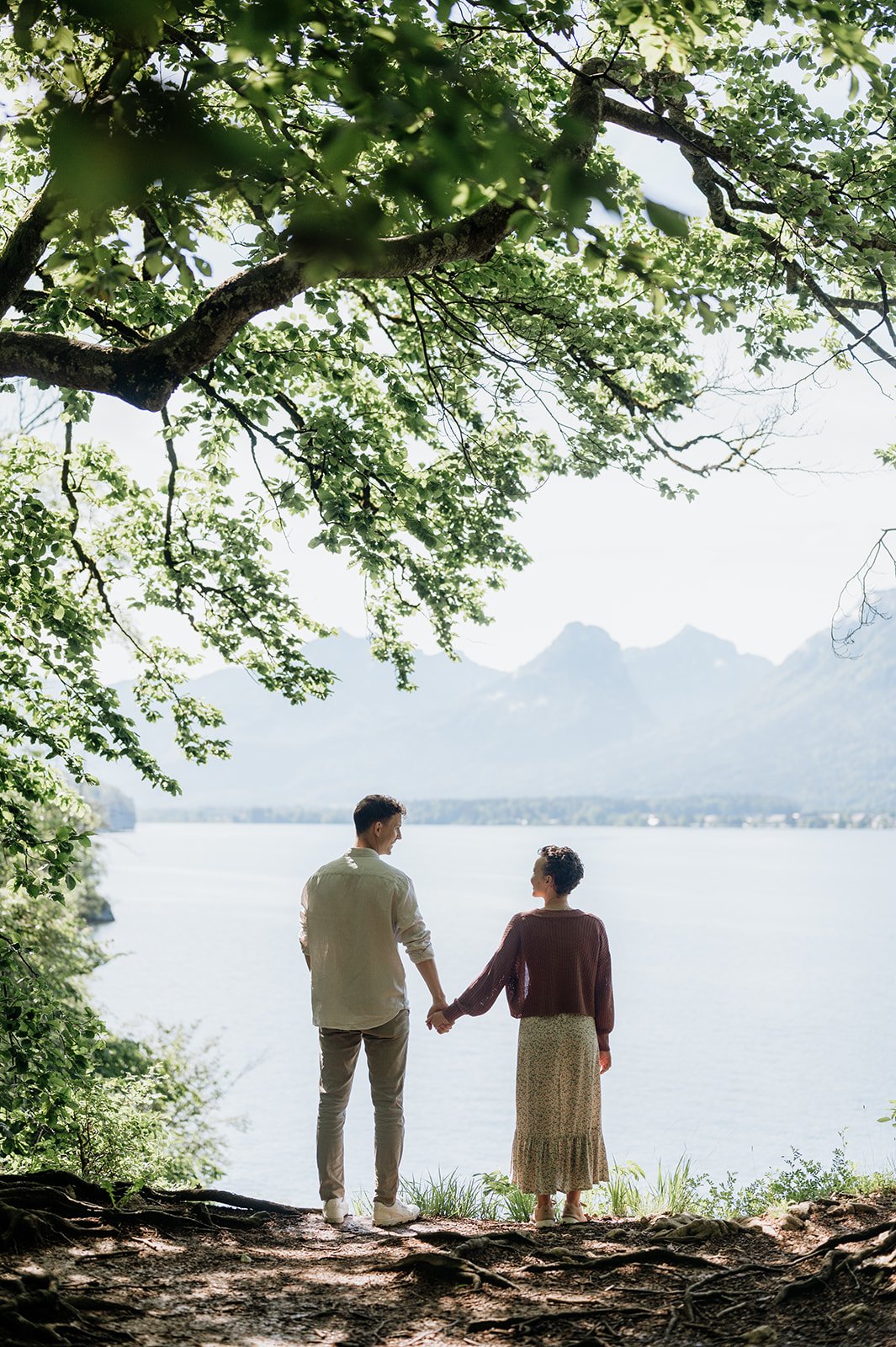 Ein Paar hält Händchen am Ufer eines Sees mit Bergen im Hintergrund, umgeben von Bäumen.