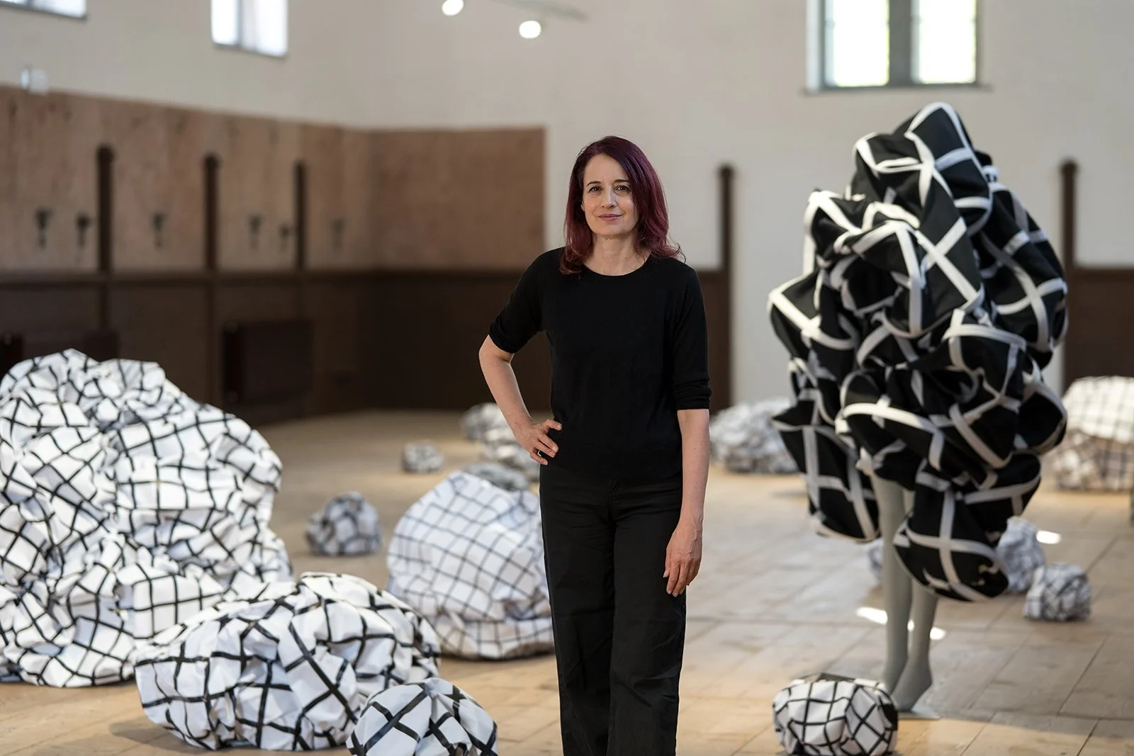Frau steht in einem Raum mit Skulpturen aus bedrucktem Stoff, die wie Papierknäuel aussehen, vor einem weißen Wandhintergrund.