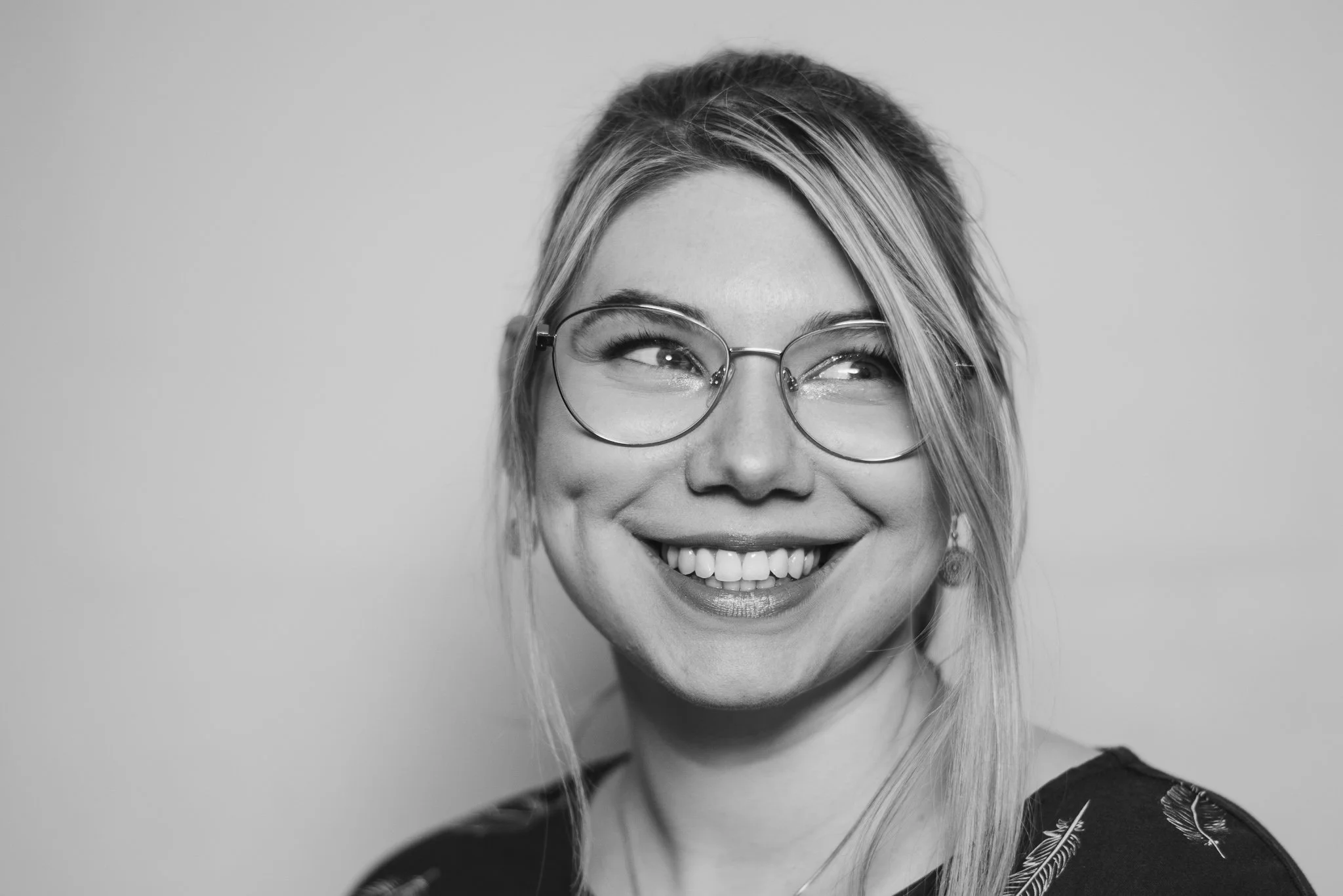 Schwarz-Weiß-Bild einer lachenden jungen Frau mit Brille, Haare zur Seite gekämmt, trägt ein dunkles Oberteil mit Feder-Muster.