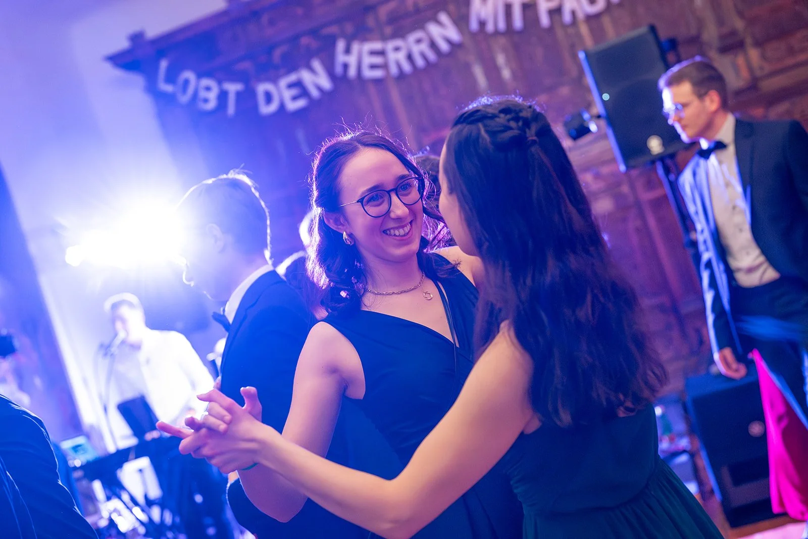 Zwei Frauen tanzen auf einer Feier im eleganten schwarzen Kleid, im Hintergrund eine Band und andere Gäste, eine Garderobe mit einer Tafelkenntnis 'Lobt den Herrn mit Pracht'.