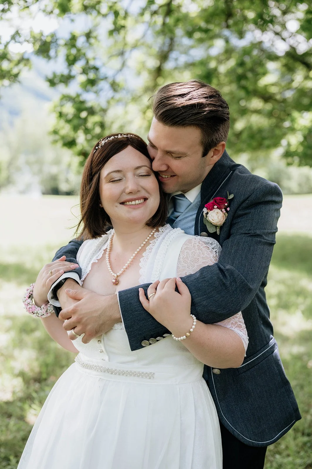 Ein Mann und eine Frau im Hochzeitsoutfit umarmen sich glücklich im Freien, umgeben von Bäumen.