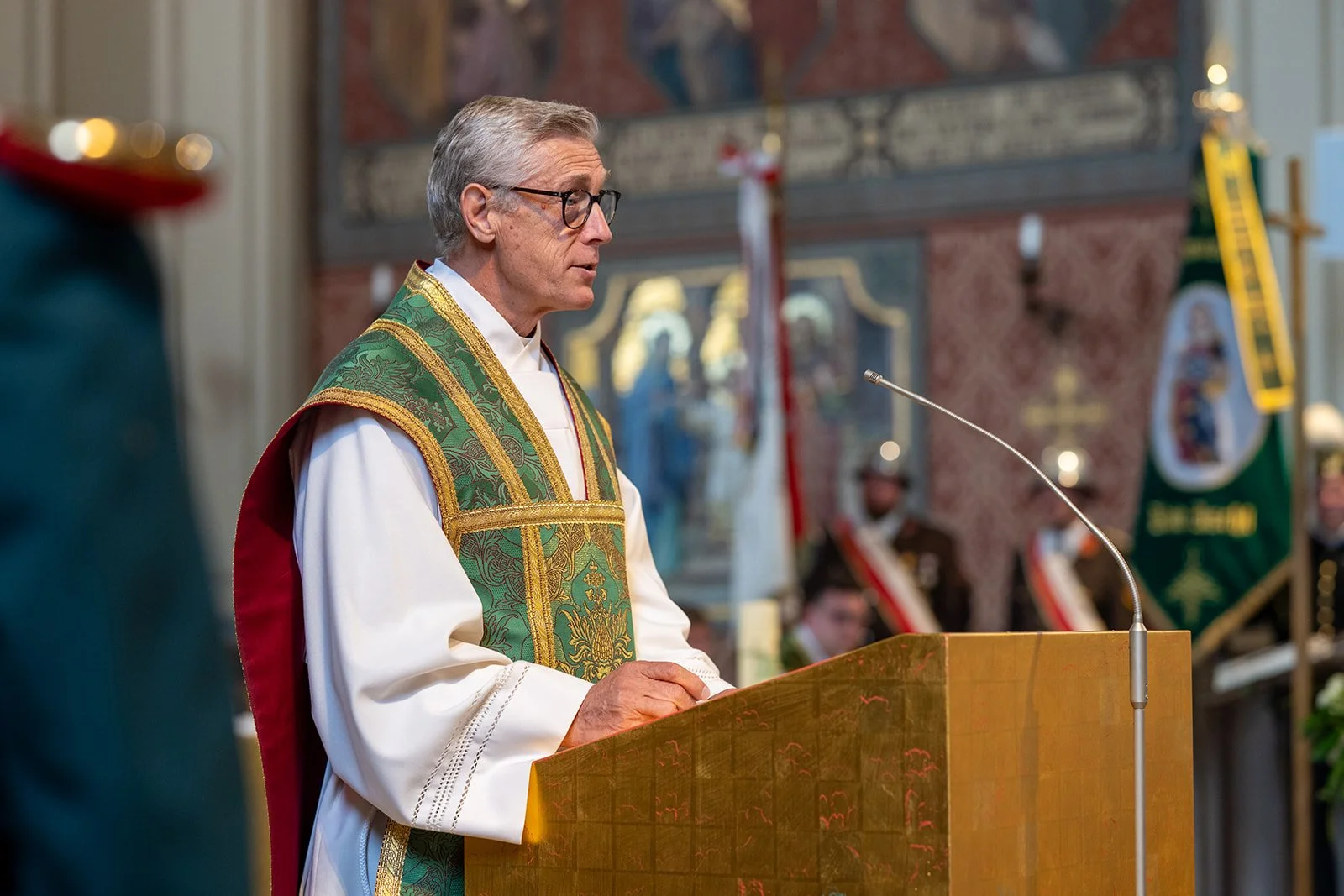 Ein Priester in liturgischer Kleidung liest während eines Gottesdienstes in einer Kirche. Im Hintergrund sind kirchliche Symbole und Flaggen sichtbar.
