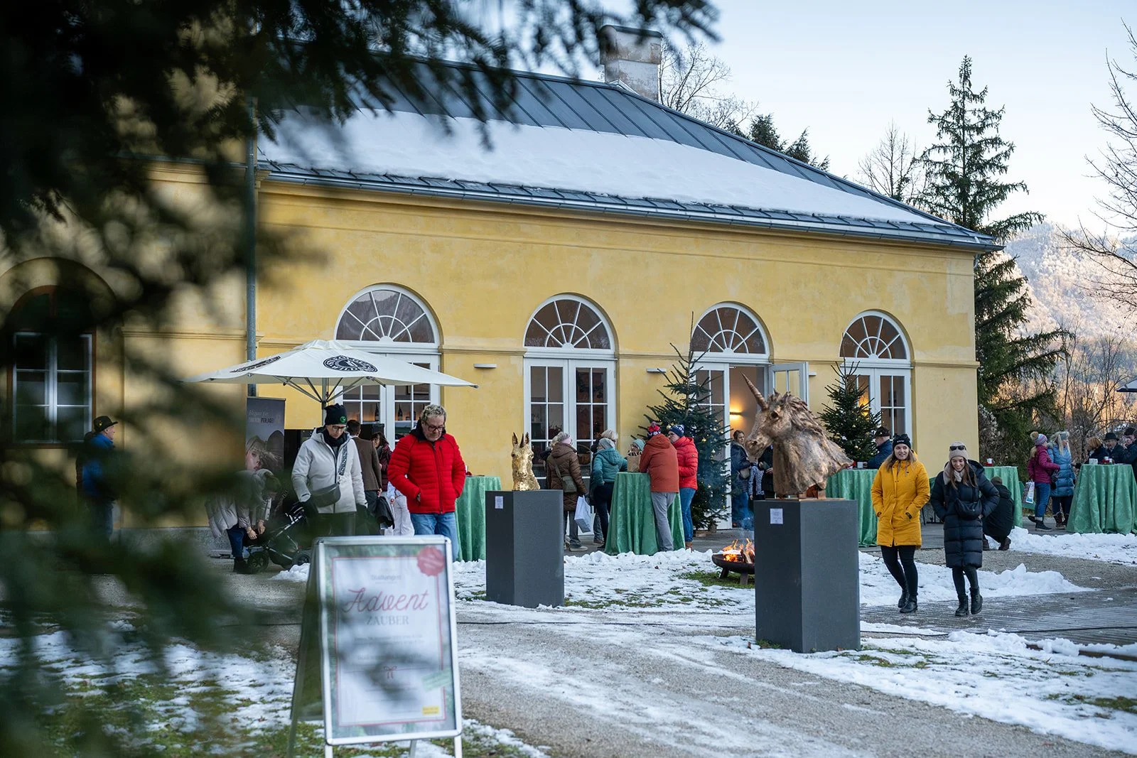 Menschen stehen während eines winterlichen Festes vor einem gelben Gebäude mit großen, halbkreisförmigen Fenstern. Es gibt Tische mit grünen Tischtüchern, Weihnachtsbäume und Tierstatuen, darunter ein Einhorn. Schnee liegt auf dem Boden und es ist ka